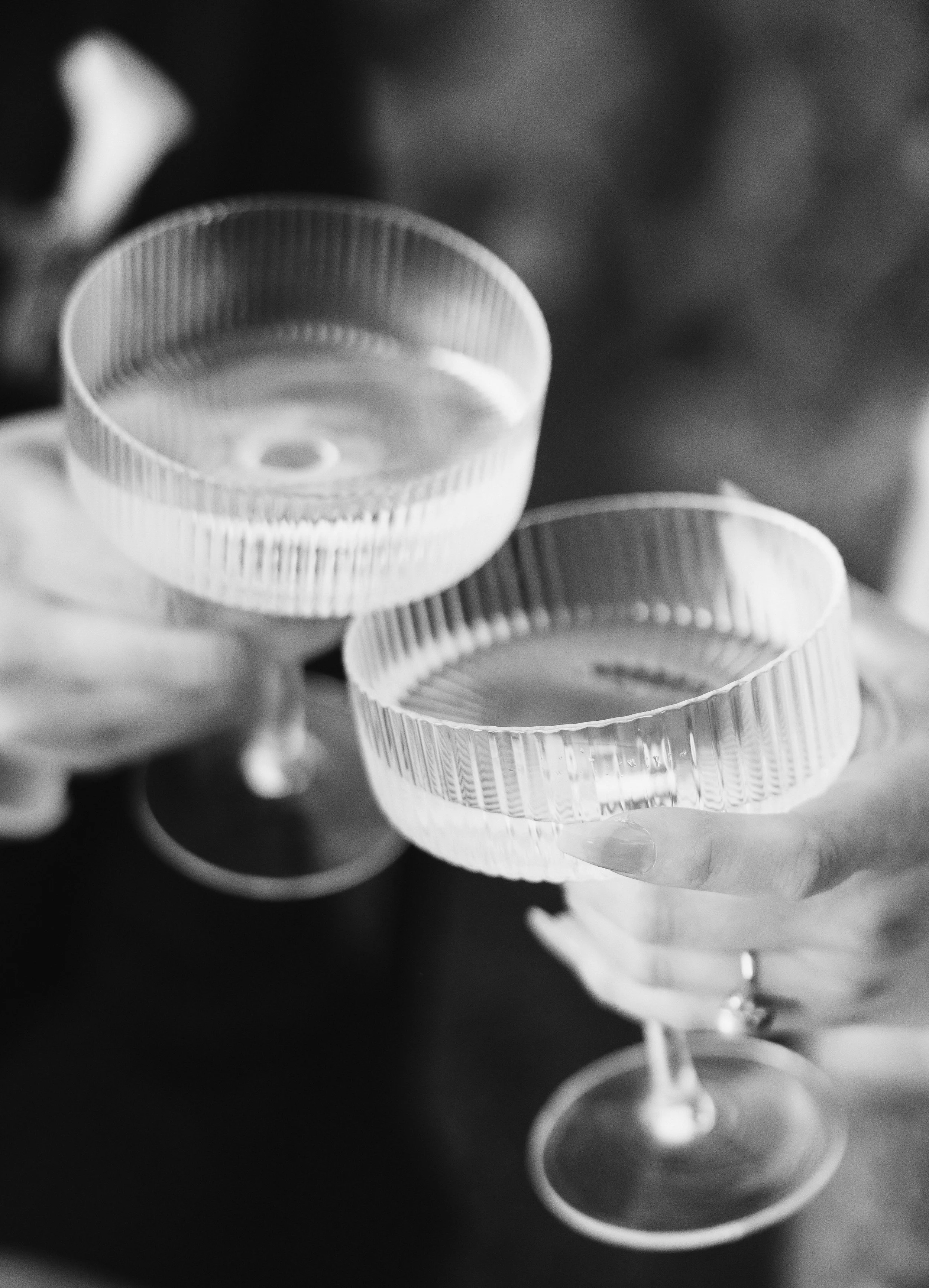Two people holding empty cocktail glasses with scalloped edges, toastings, in black and white photograph.