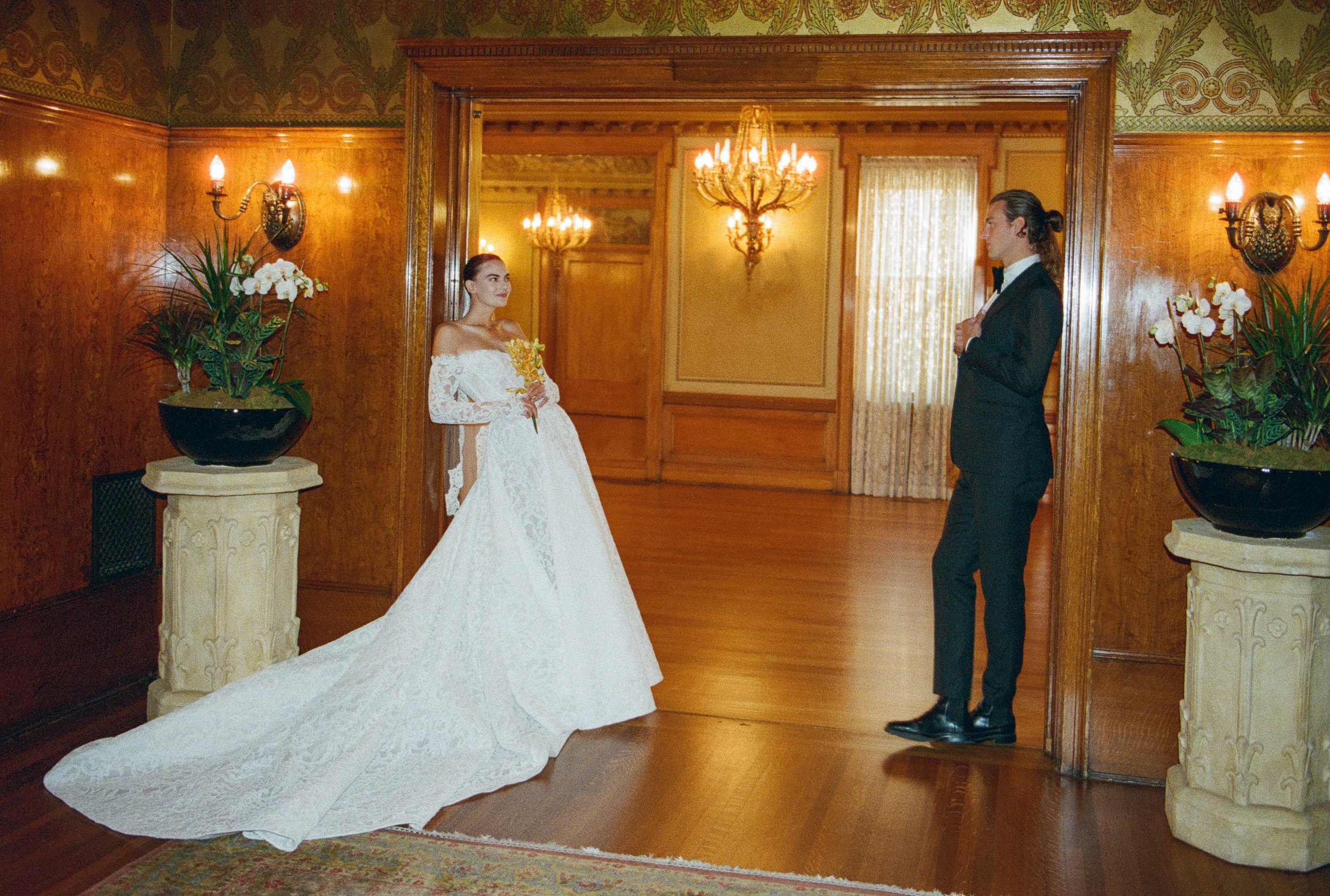 A bride in a white lace wedding gown holding a bouquet looks at a groom in a black tuxedo in a vintage decorated room with wood-paneled walls, gold chandeliers, and floral arrangements.