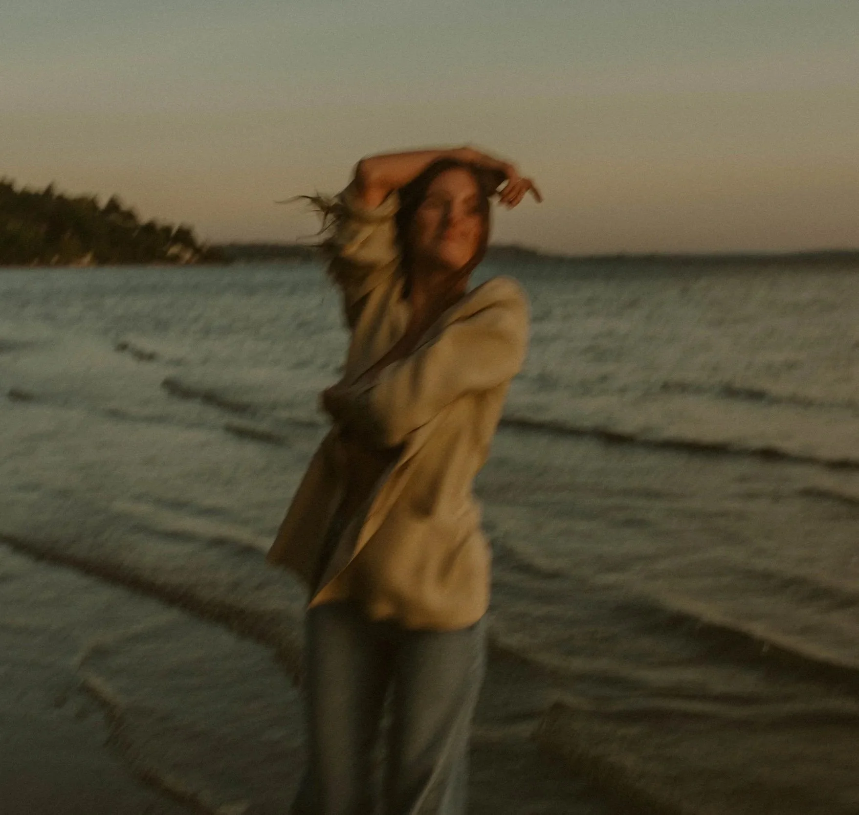 A woman standing on the beach during sunset, with her hand on her head, wearing a tan jacket and gray pants.
