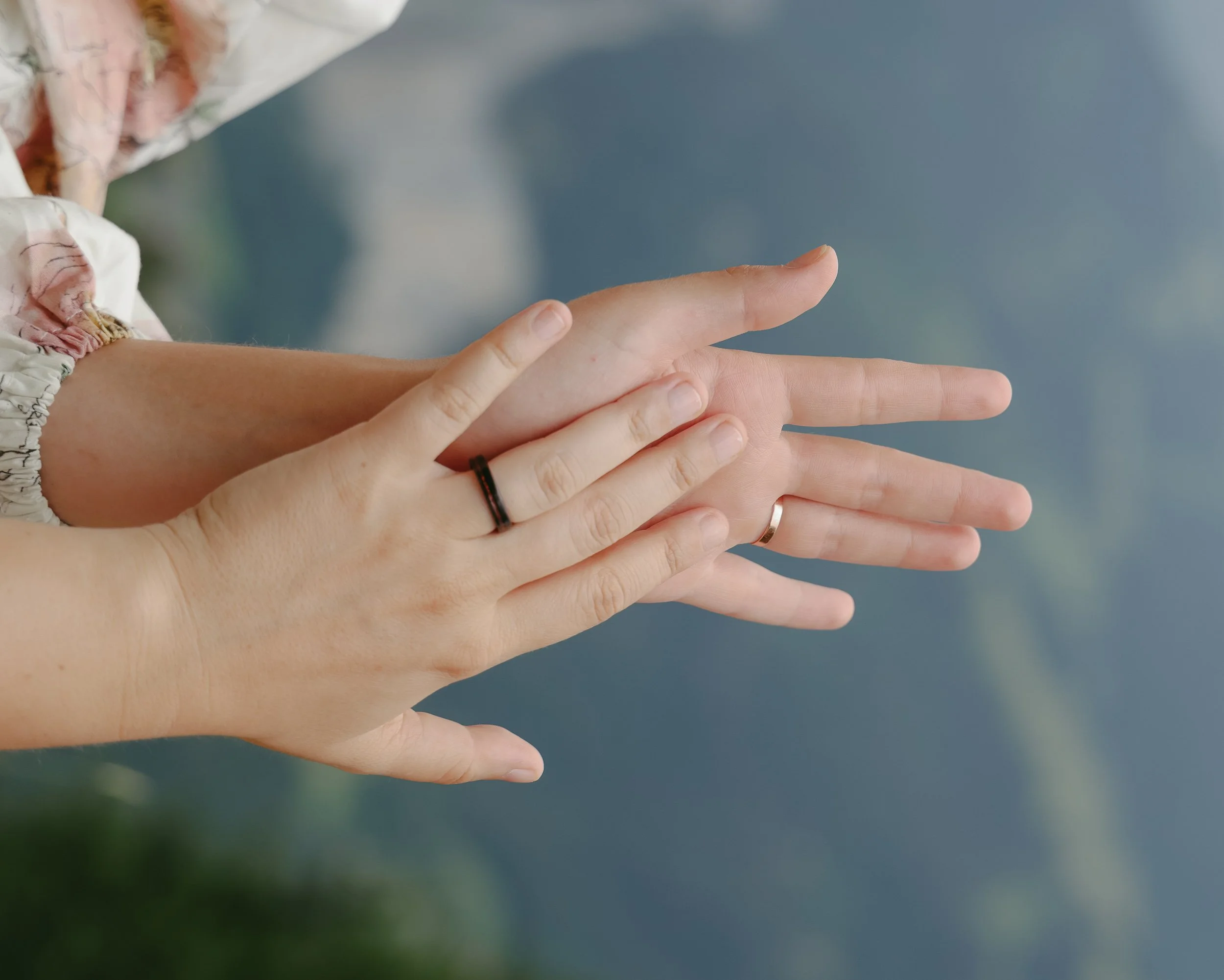 Two hands overlapping, one with a black ring and the other with a silver ring, against a blurred background of water and landscape.