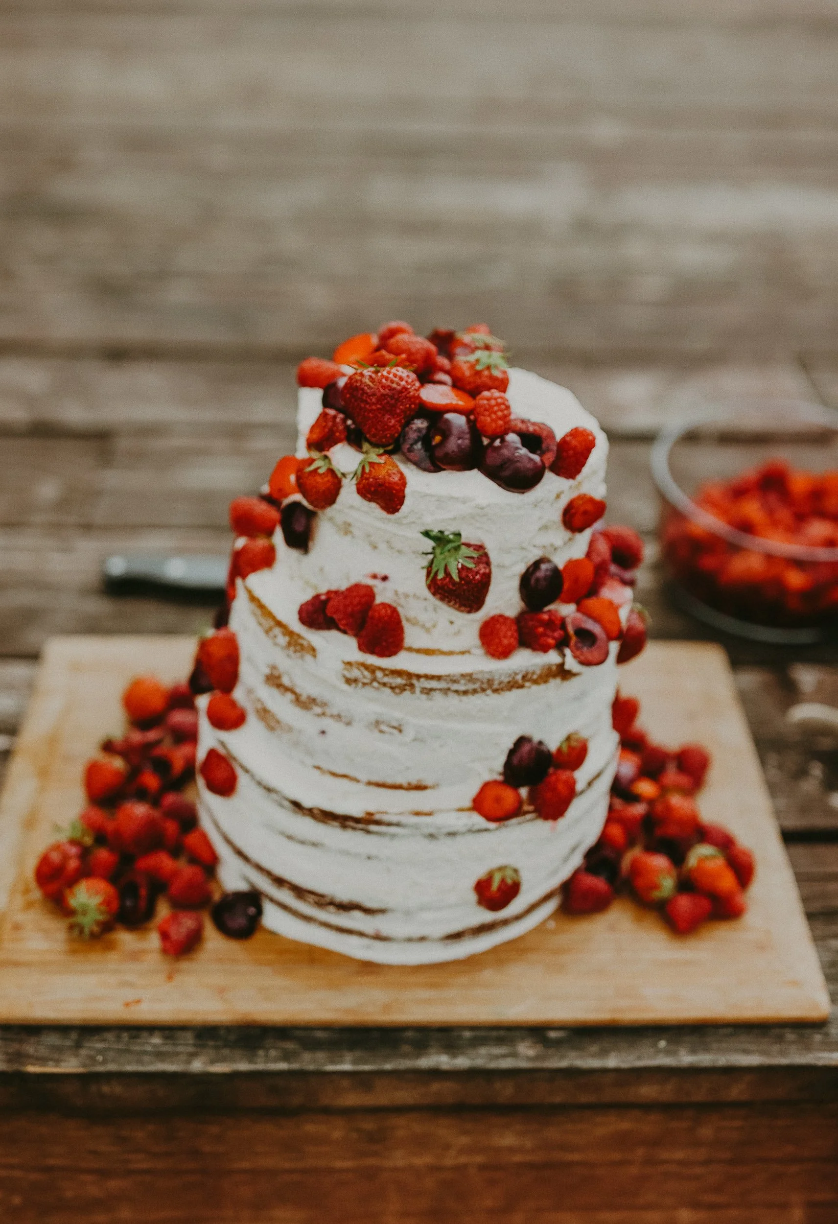A multi-layered naked cake topped with fresh berries including strawberries, raspberries, and blueberries, with additional berries scattered around the cake on a wooden surface.