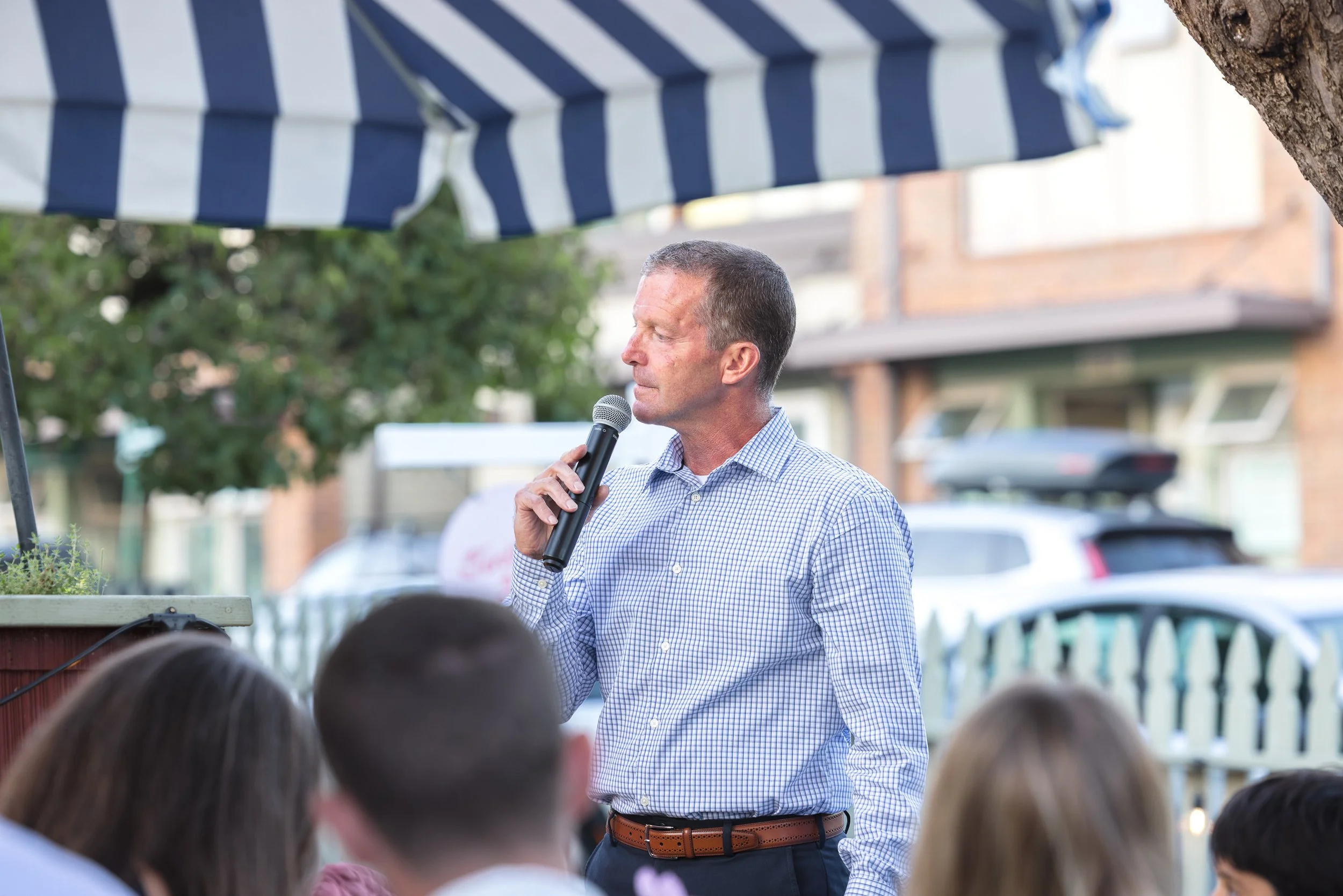 A man in a checkered shirt holding a microphone and speaking at an outdoor gathering with an audience, under a canopy with blue and white stripes.