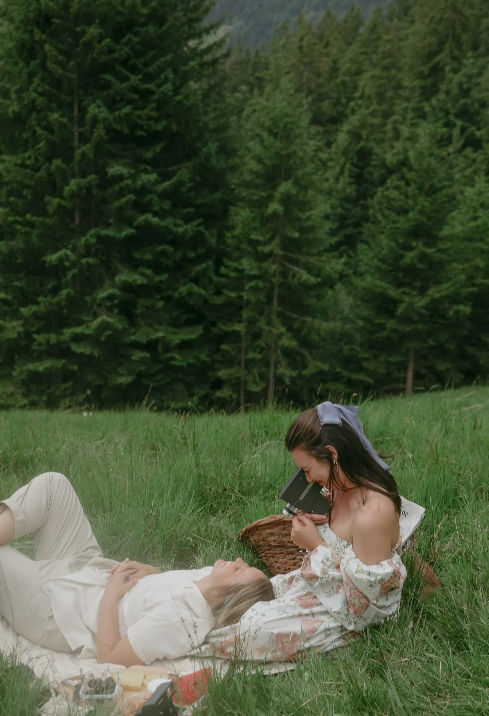 Two women in a grassy field with tall trees in the background. One woman is lying on her back, while the other is sitting and looking through a vintage camera.