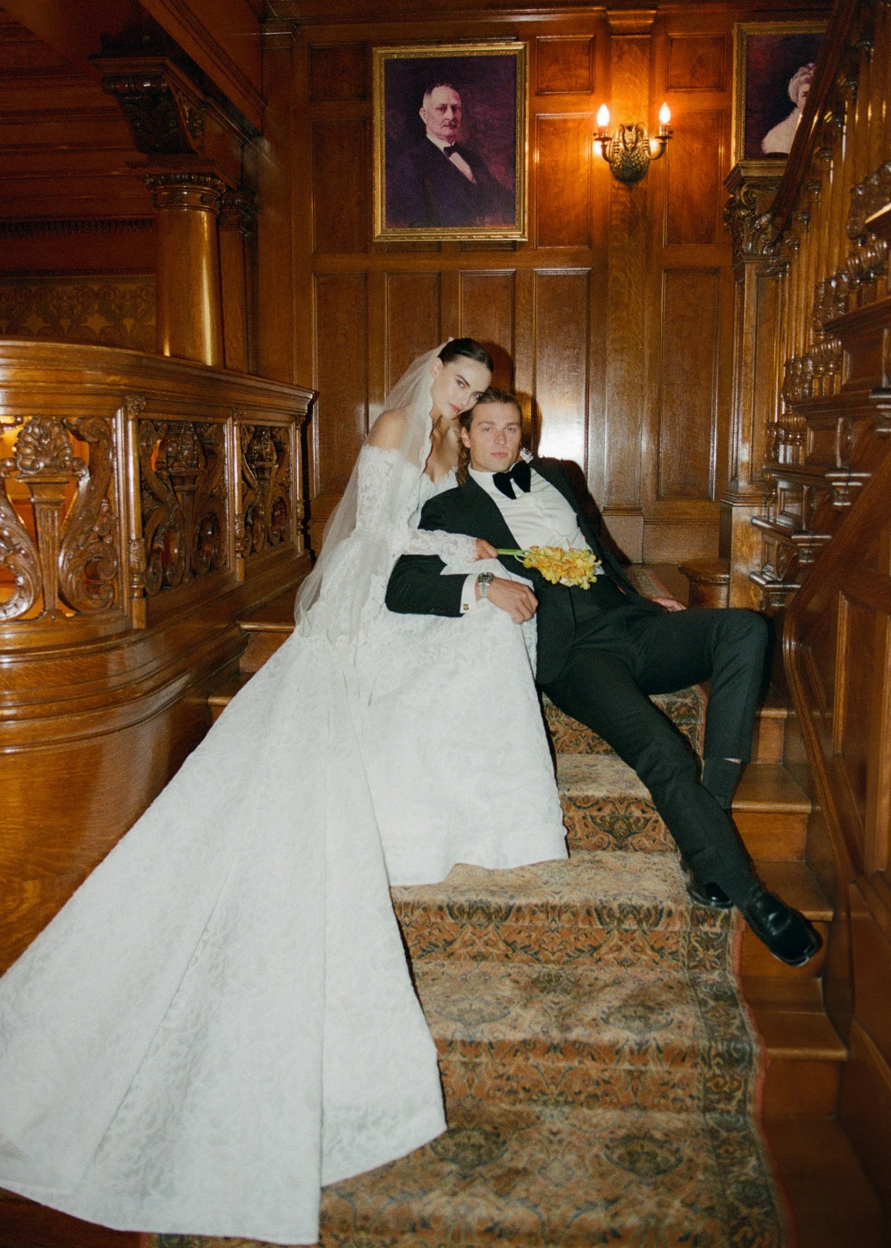A bride and groom in formal wedding attire sitting on a staircase inside a room with wood-paneled walls and portraits hanging on the wall.