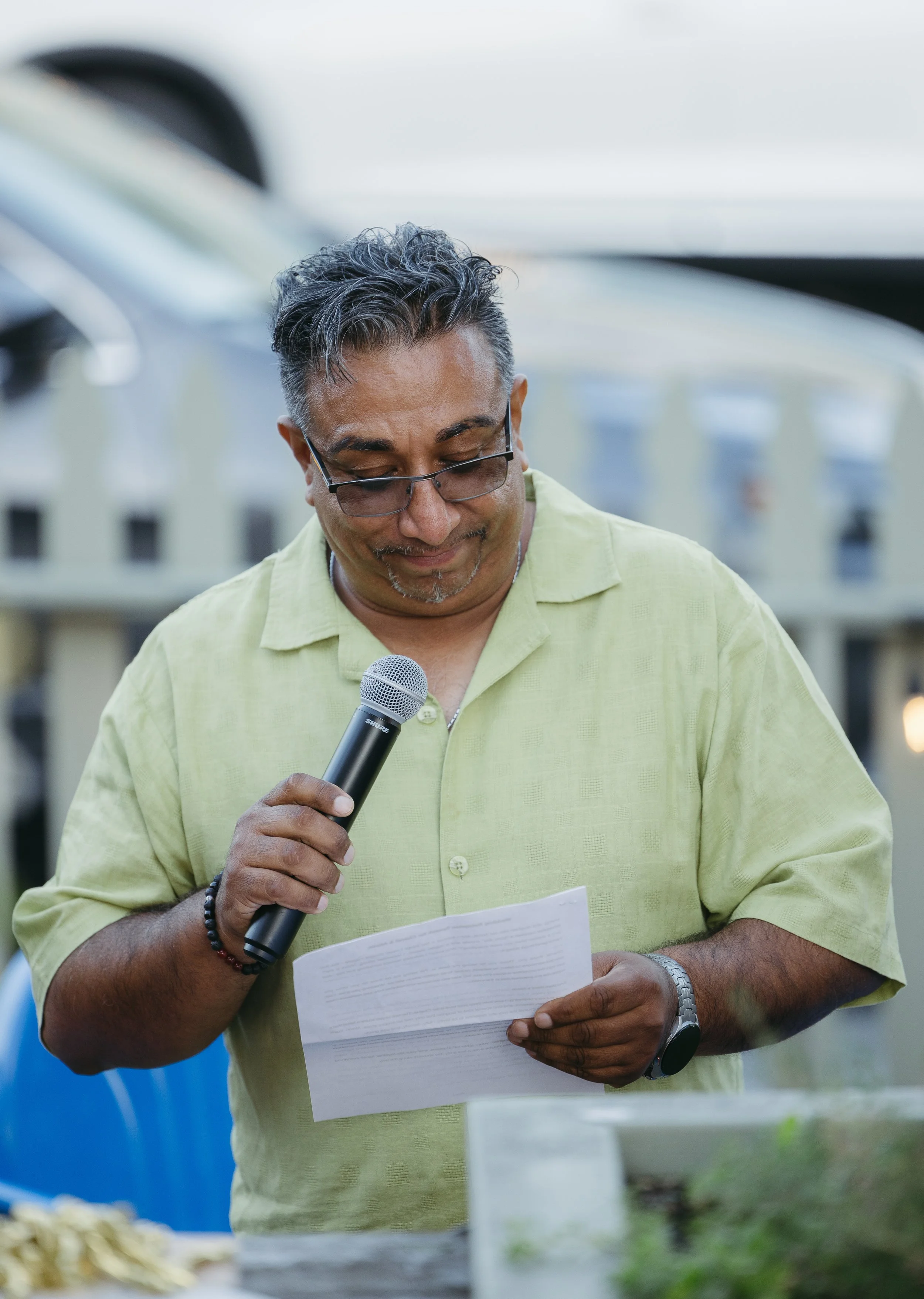 A man with glasses and a gray beard wearing a light green shirt, holding a microphone in his right hand and a paper in his left hand, standing outdoors with a blurred background.