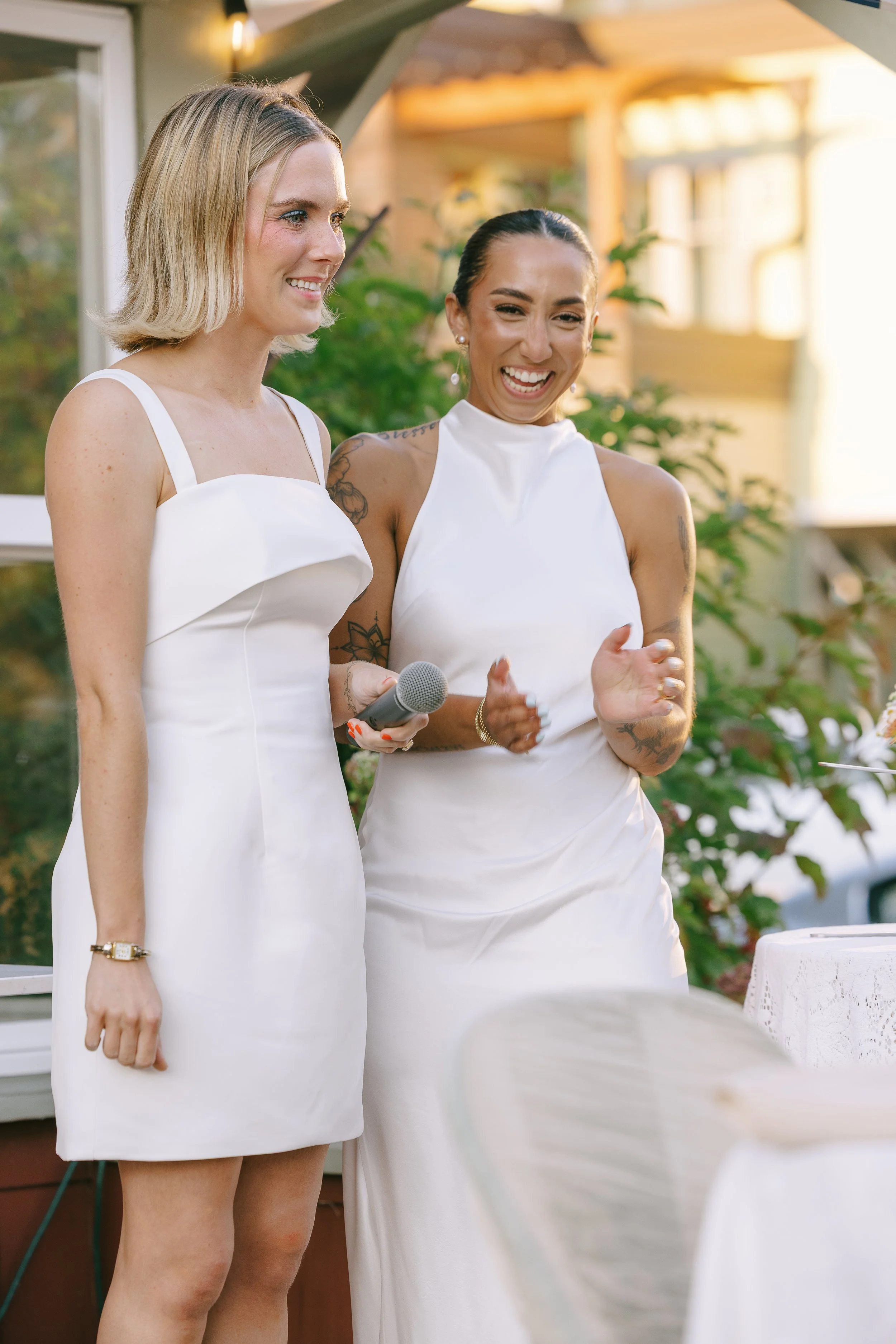 Two women in white dresses smiling and clapping at an outdoor event, one holding a microphone.