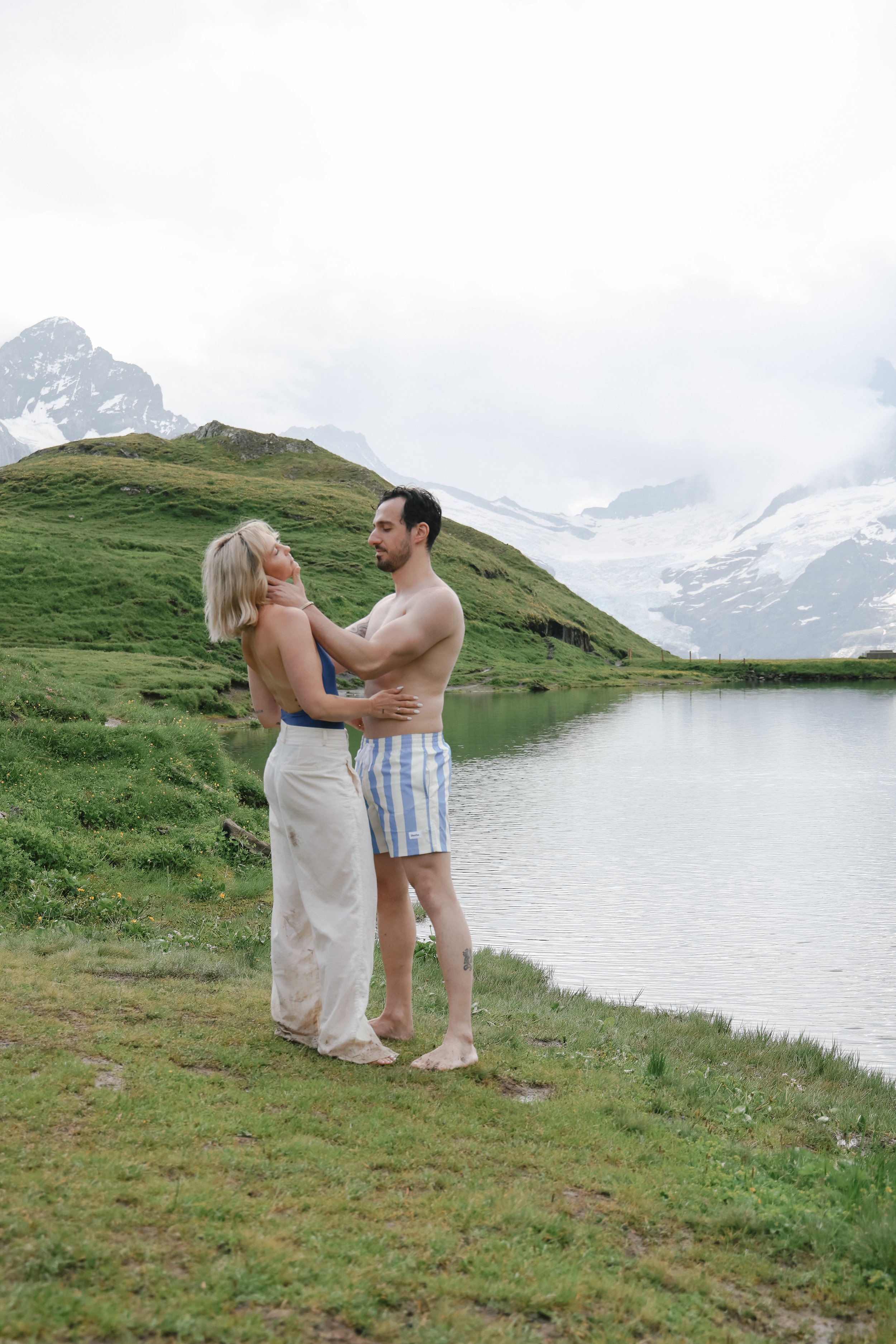 A couple stands close by a lake with green hills and snow-capped mountains in the background. The woman is in a long beige skirt and blue top, while the man is in striped shorts. They are holding each other and gazing into each other's eyes against a