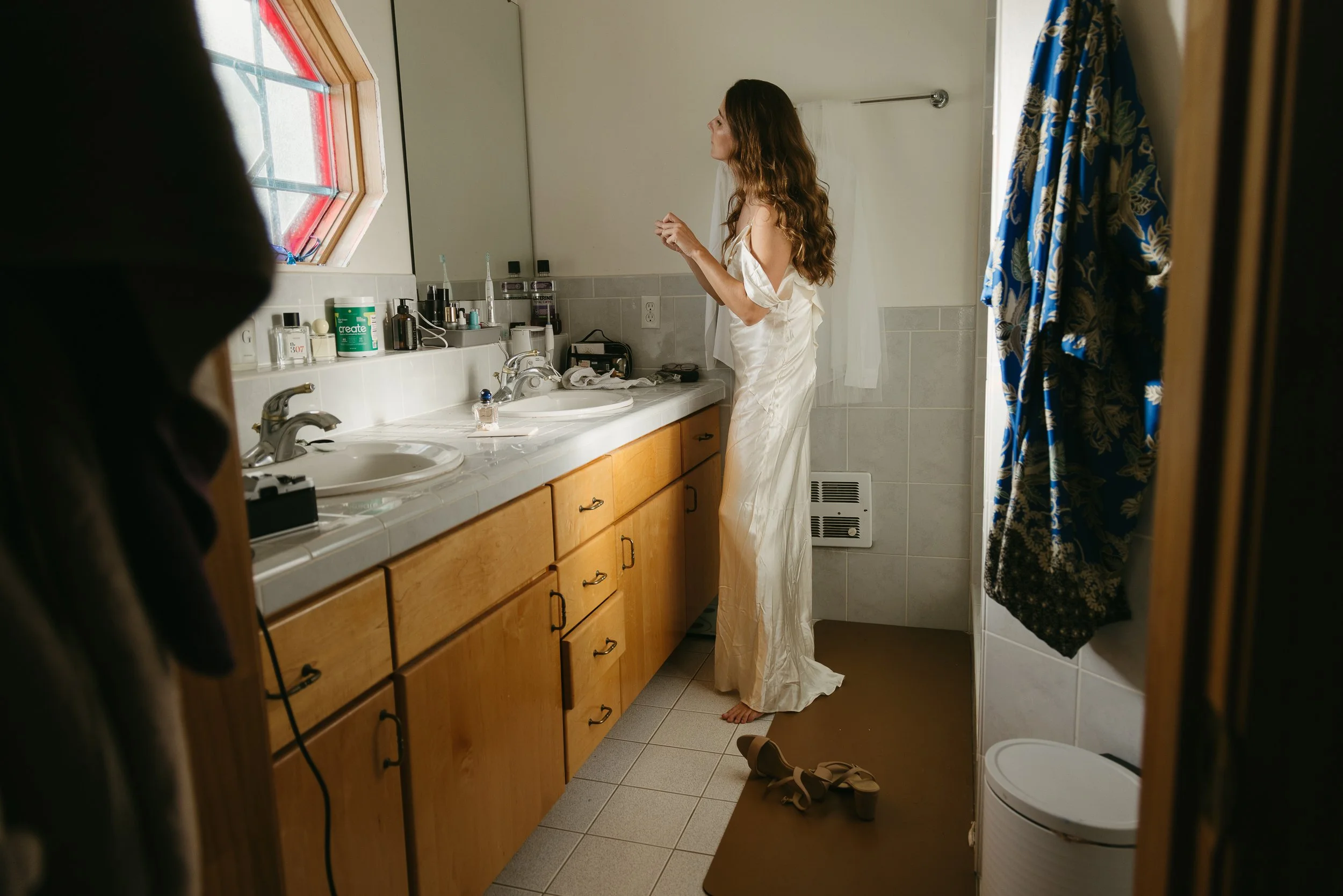 A woman standing in a bathroom at a double sink vanity, preparing or adjusting her hair or makeup, wearing a silk dress. The bathroom has a window, various toiletries on the counter, a blue patterned shower curtain, and some shoes on a mat.