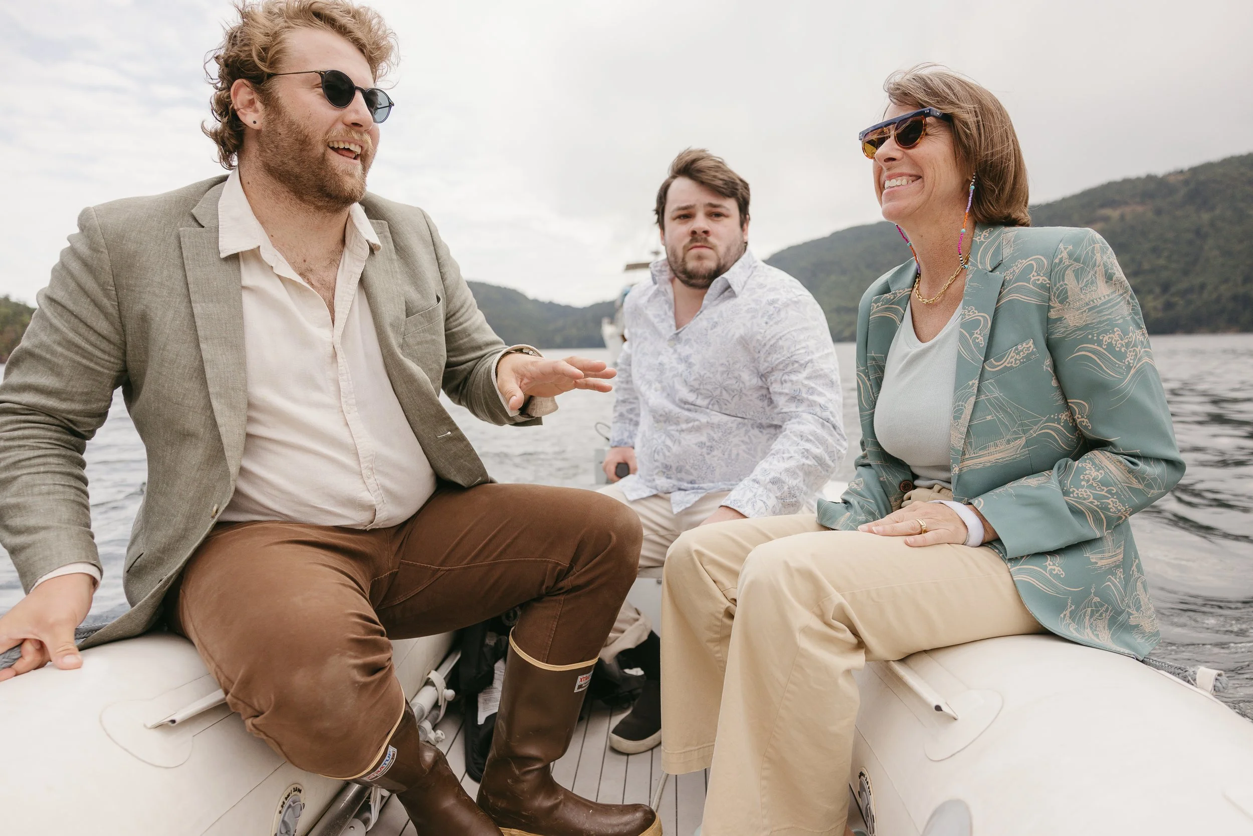Three people sitting in an inflatable boat on a lake, talking and smiling, with hills in the background.