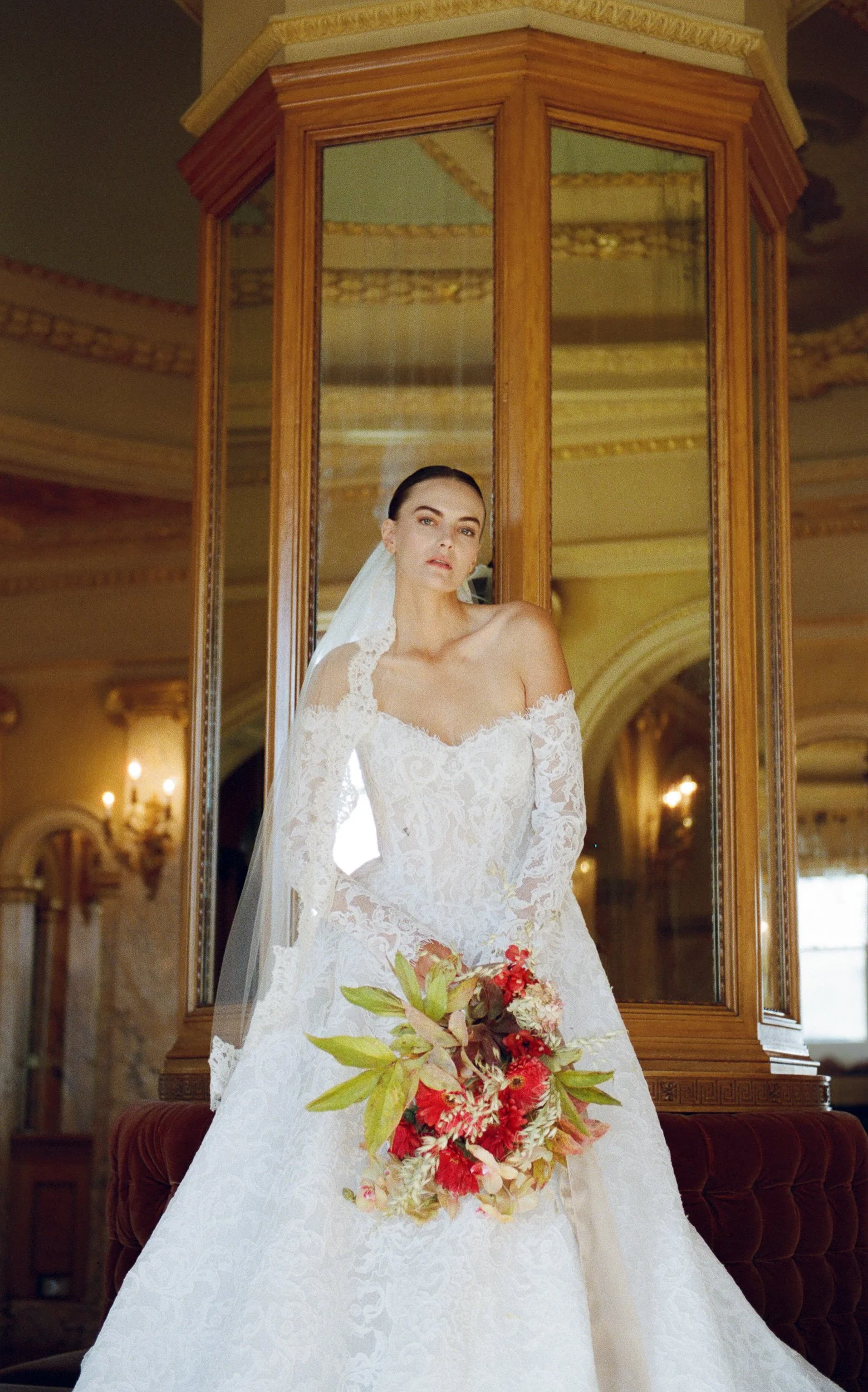 Bride in a white lace wedding gown with an off-the-shoulder neckline, holding a bouquet of red and pink flowers with green leaves, standing in front of a glass cabinet in an ornate room.