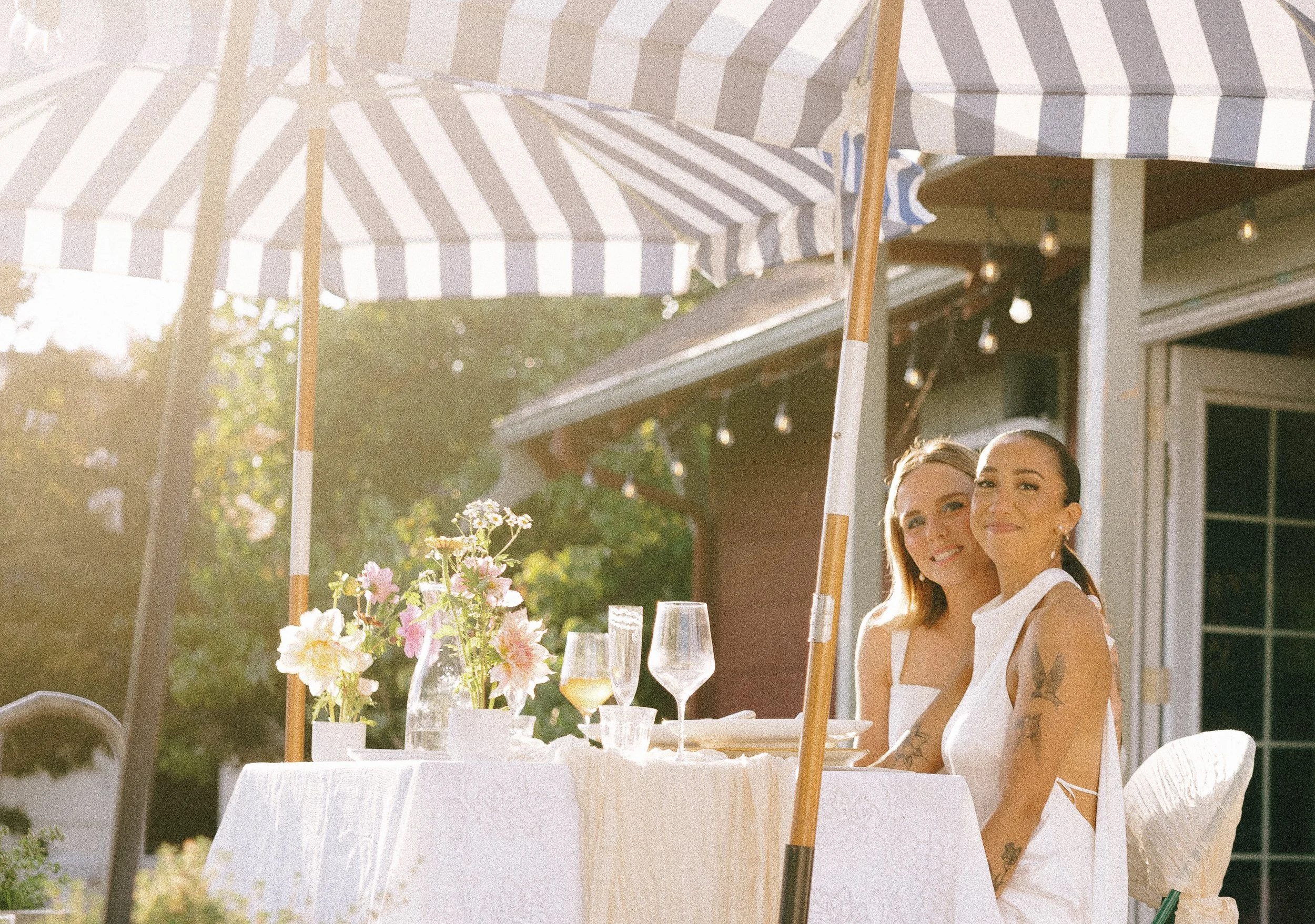 Two women sitting at a table outdoors under a striped patio umbrella, smiling. The table has a white tablecloth, a vase with pink and white flowers, and several glasses of wine. The background shows a house and trees.
