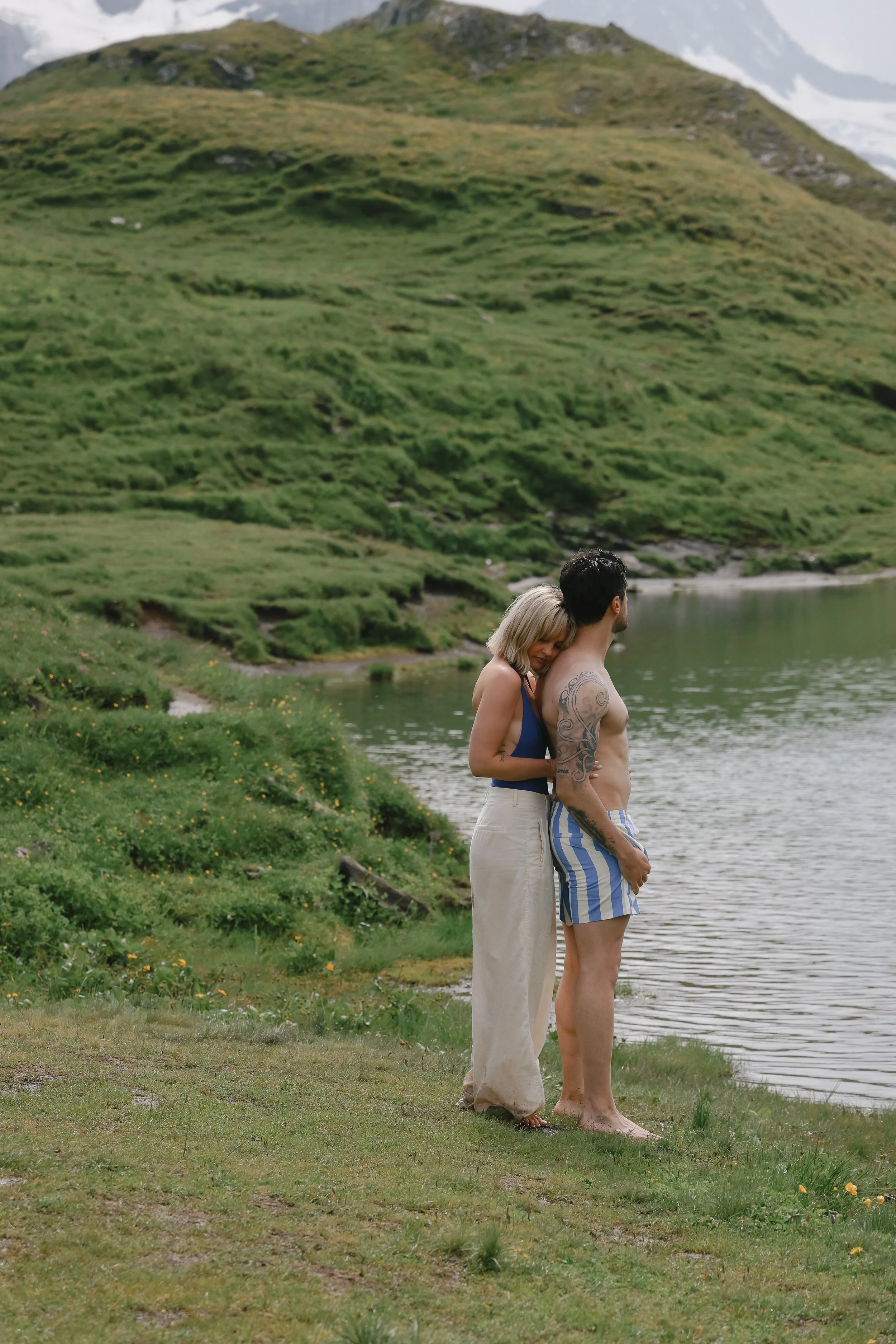 A man and woman standing close by a lake in a green, hilly landscape. The woman is resting her head on the man's shoulder.