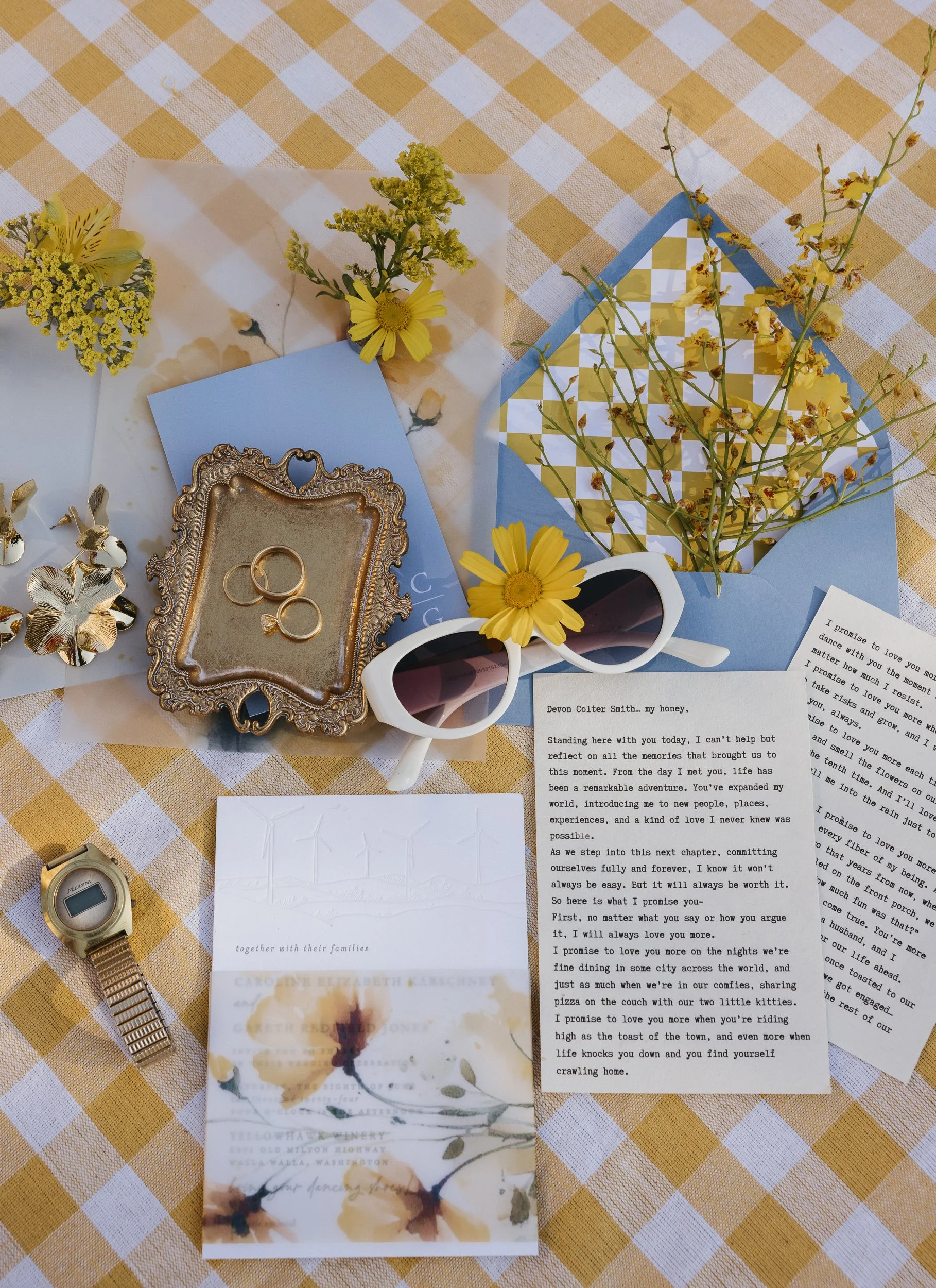 A flat lay of a table with yellow and white checkered tablecloth, featuring flowers, a gold ring box with rings, a wristwatch, sunglasses with a yellow flower, and handwritten and printed letters.