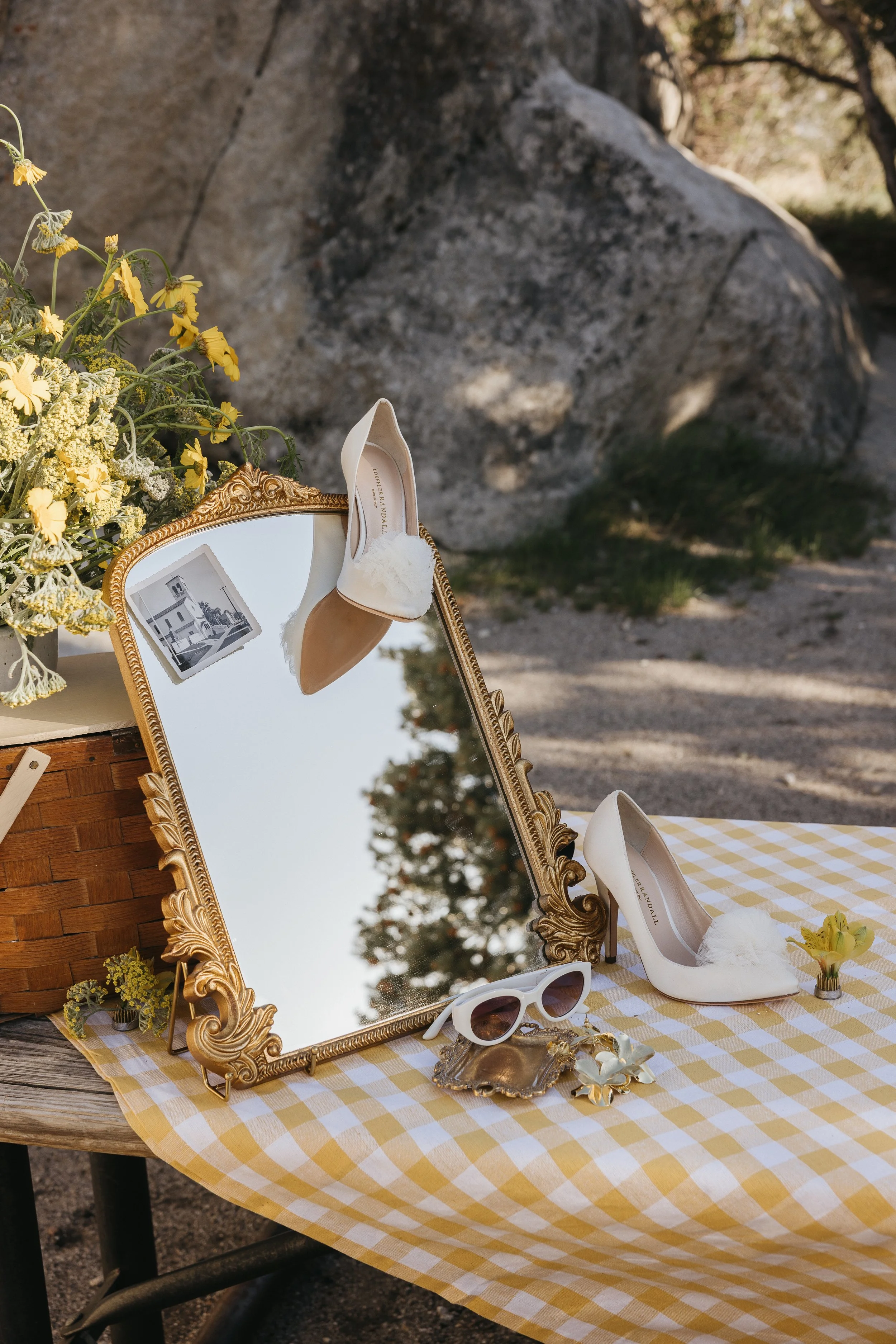 A vintage mirror leaning against a wooden table, with a pair of white wedding shoes resting on top of it. On the table are sunglasses, a small decorative box, some jewelry, a checkered yellow and white cloth, a small yellow flower, and a flower arrangement with yellow flowers. The scene is outdoors with rocks and trees in the background.