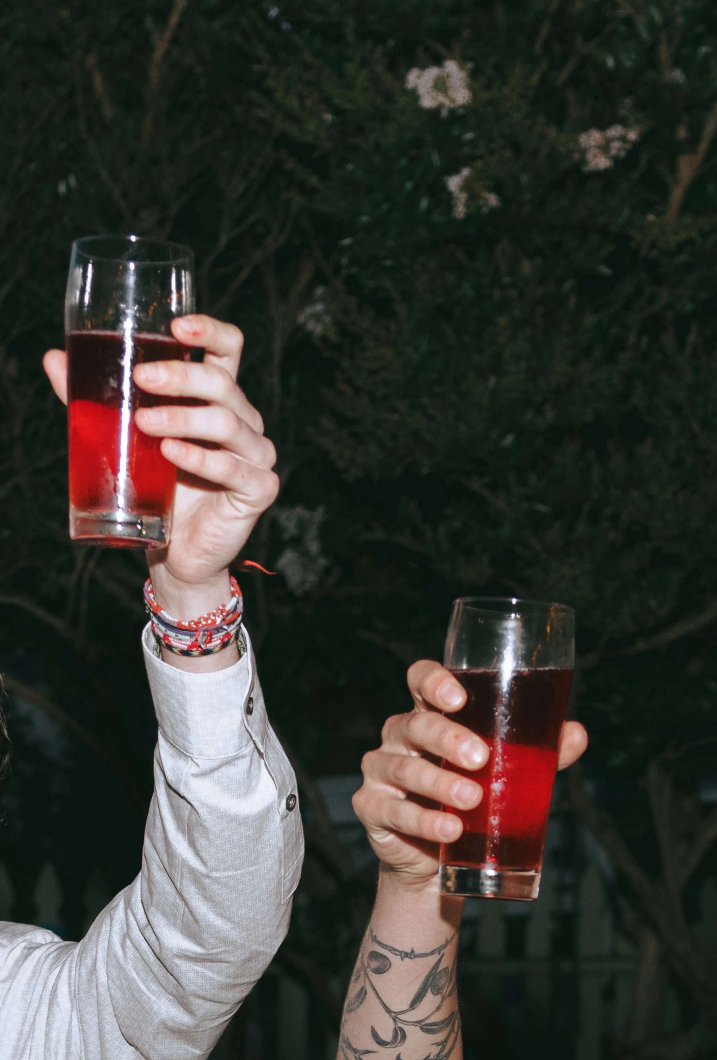 Two people holding glasses of red beverage outdoors at night, one with a sleeve and bracelet, the other with a tattooed arm.