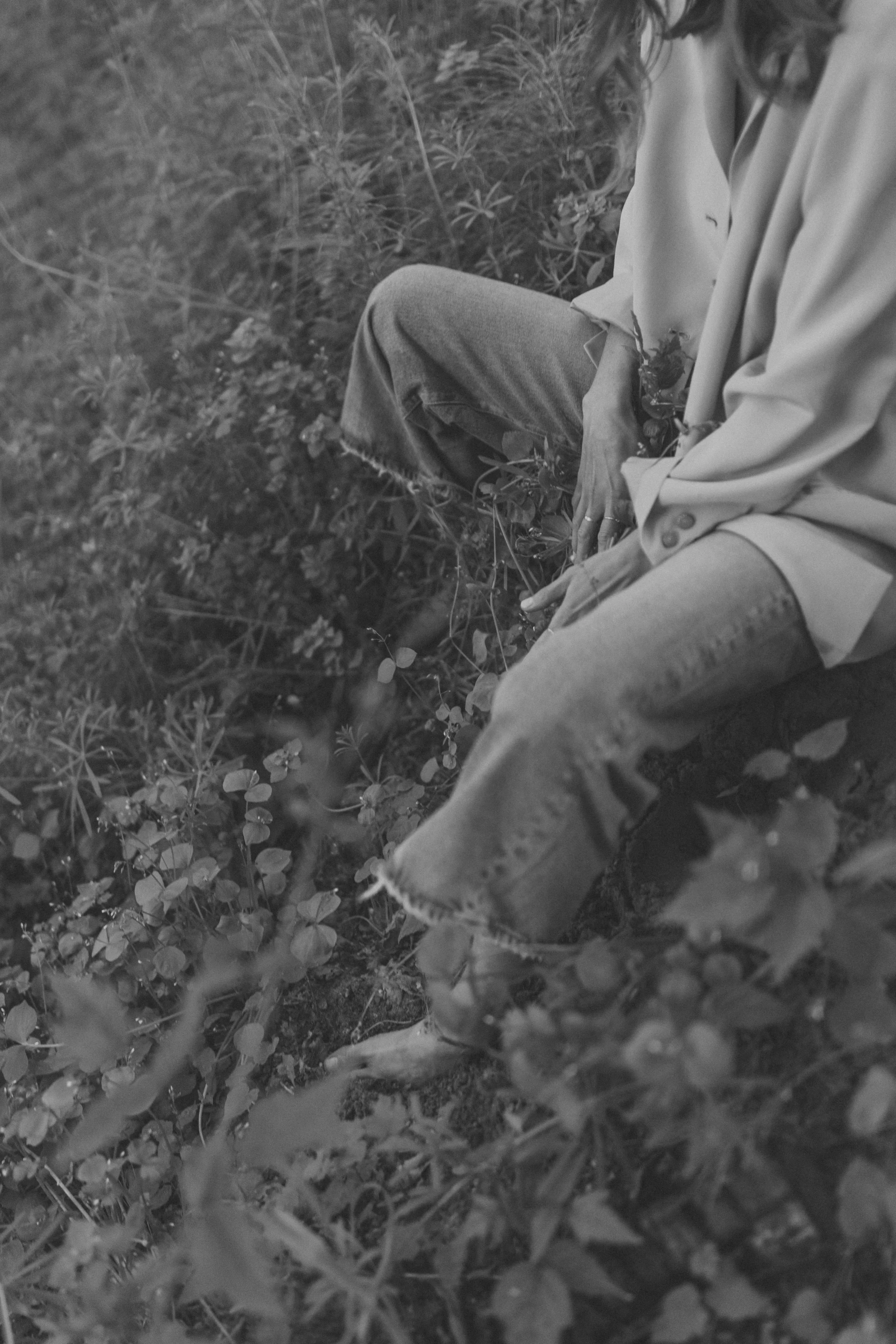 A person sitting among low plants and grass, wearing ripped jeans and a long-sleeved shirt, with their face not visible.