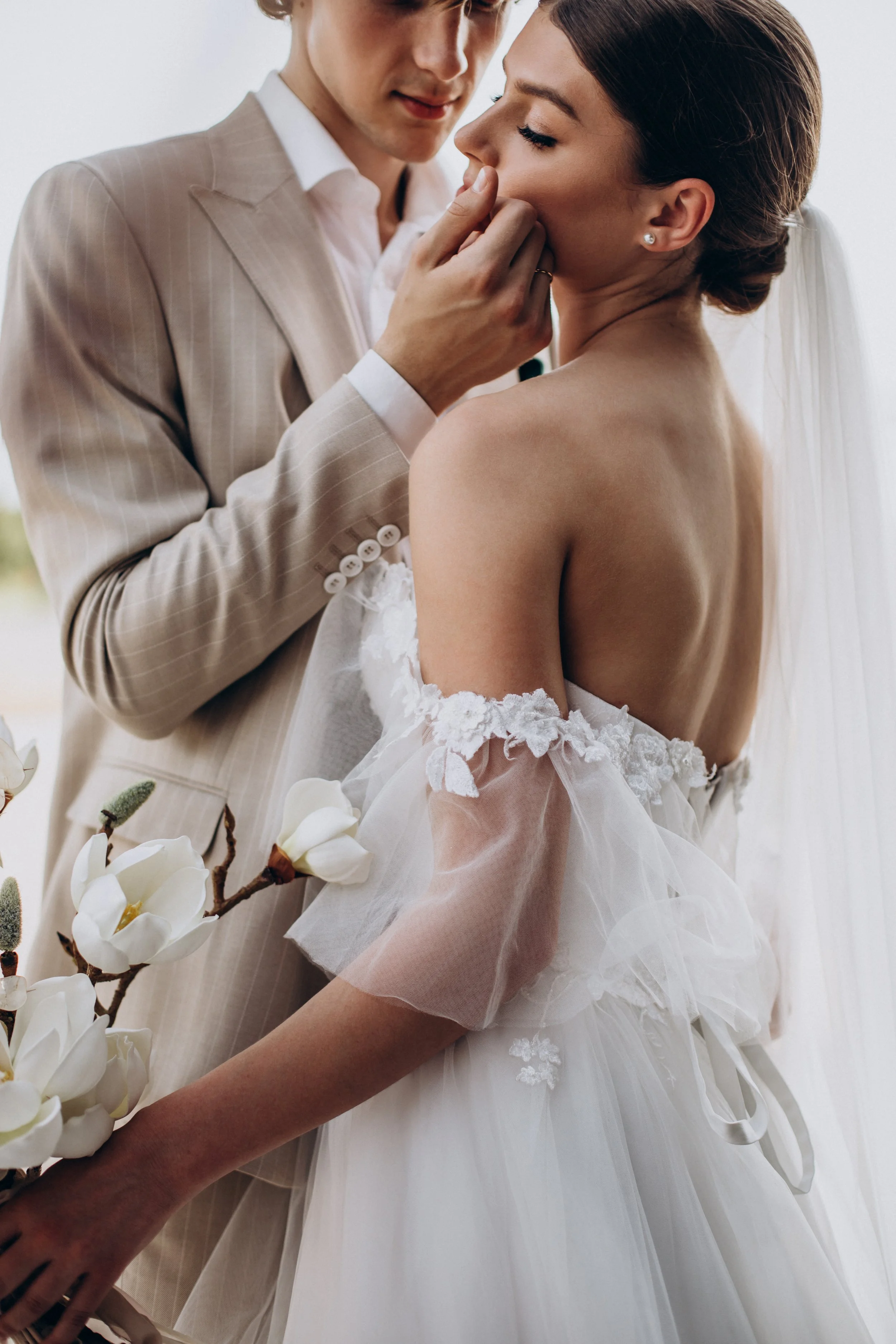 A wedding scene with a groom gently touching the bride's face, who is wearing a white wedding dress with off-shoulder lace and a veil, holding a white flower bouquet.