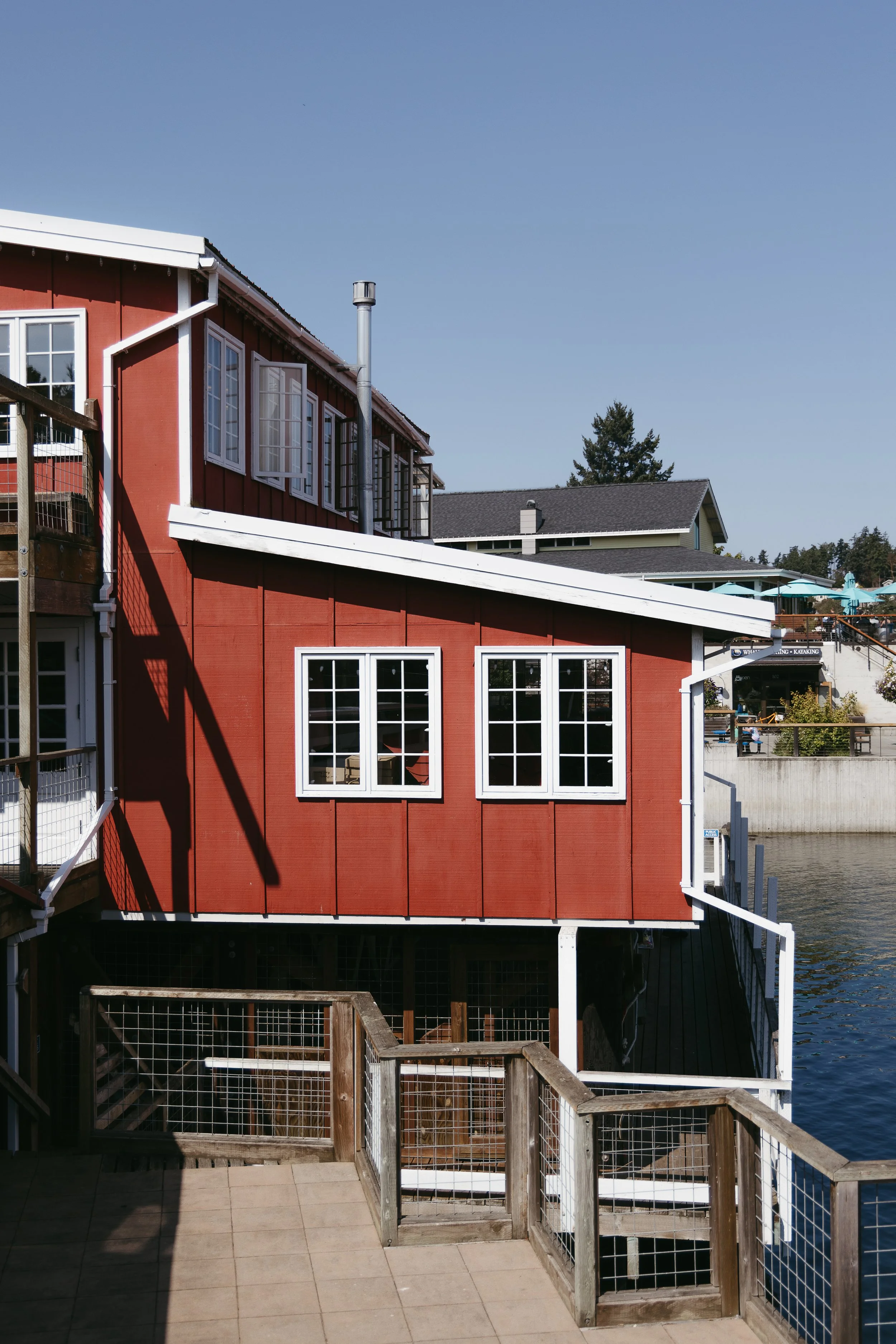 Red building by the water with white-framed windows and a sloped roof, seen from an outdoor tiled balcony with wooden railings.