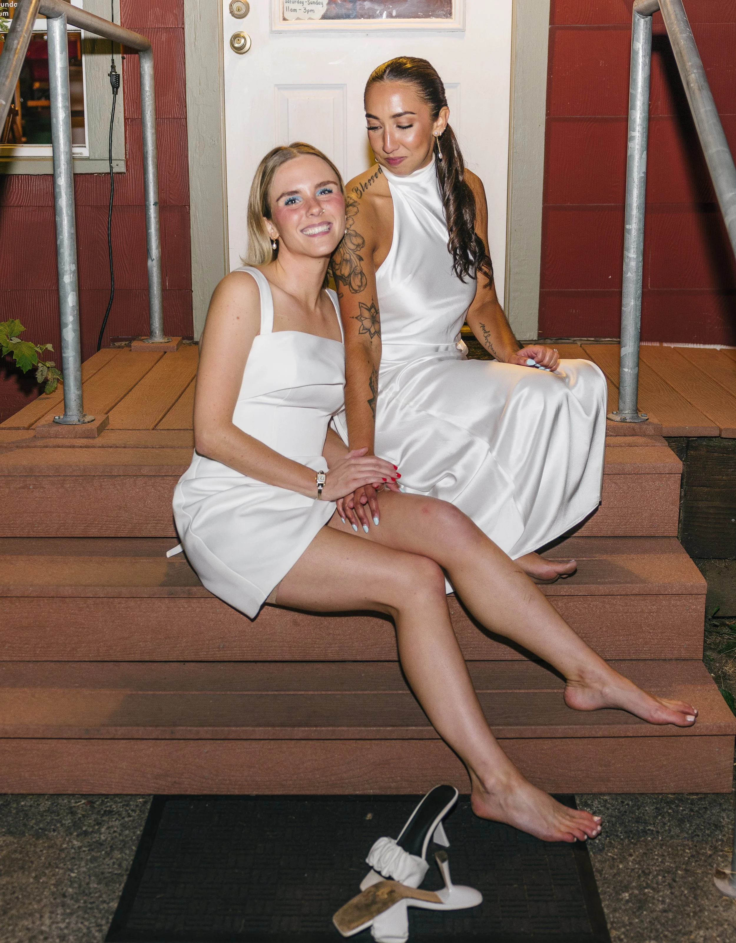 Two women wearing white dresses sitting on wooden steps outside a building, smiling and posing for the camera.