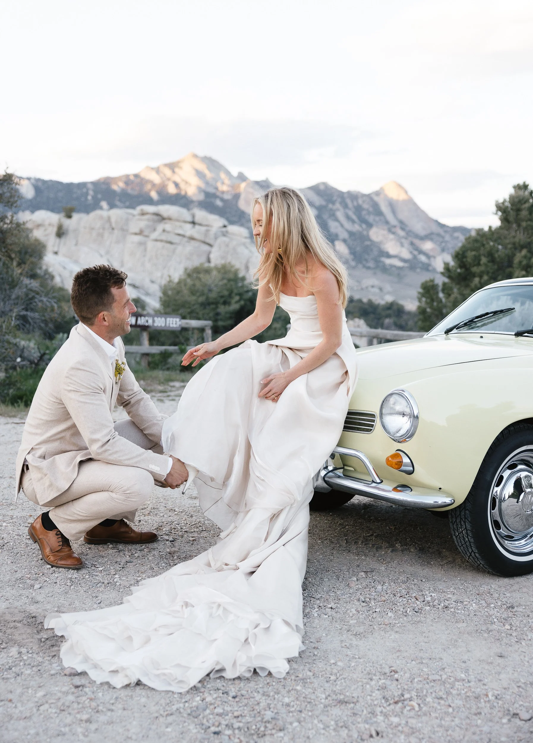 A man kneeling on the ground holding a woman’s leg, sitting on a vintage yellow car, in an outdoor mountainous setting, dressed in wedding attire.