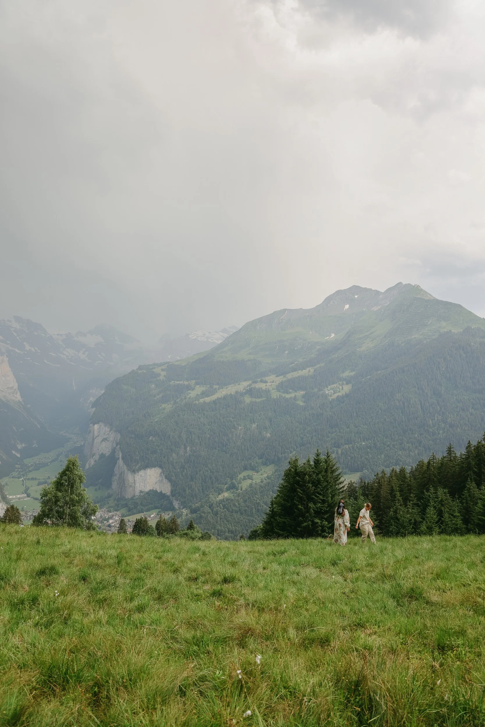 Two people walking on a grassy field with a backdrop of mountains and forest under a cloudy sky.