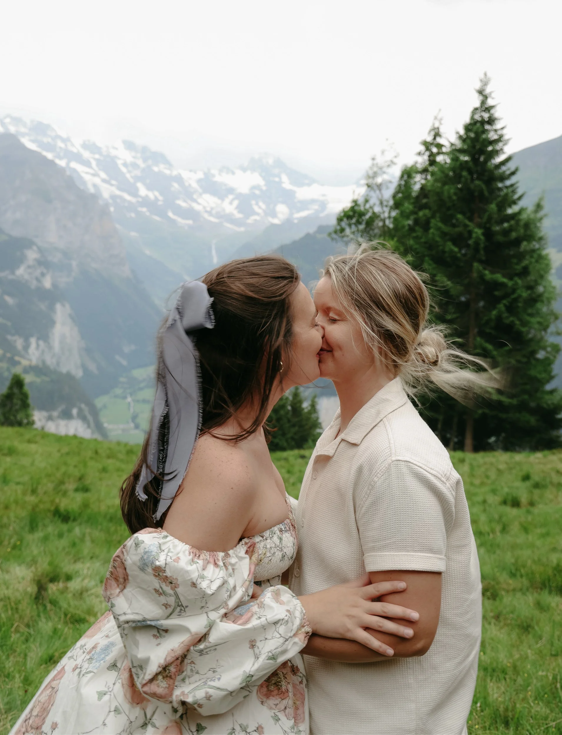 Two women kissing in a scenic mountain landscape with snow-capped peaks and tall pine trees.