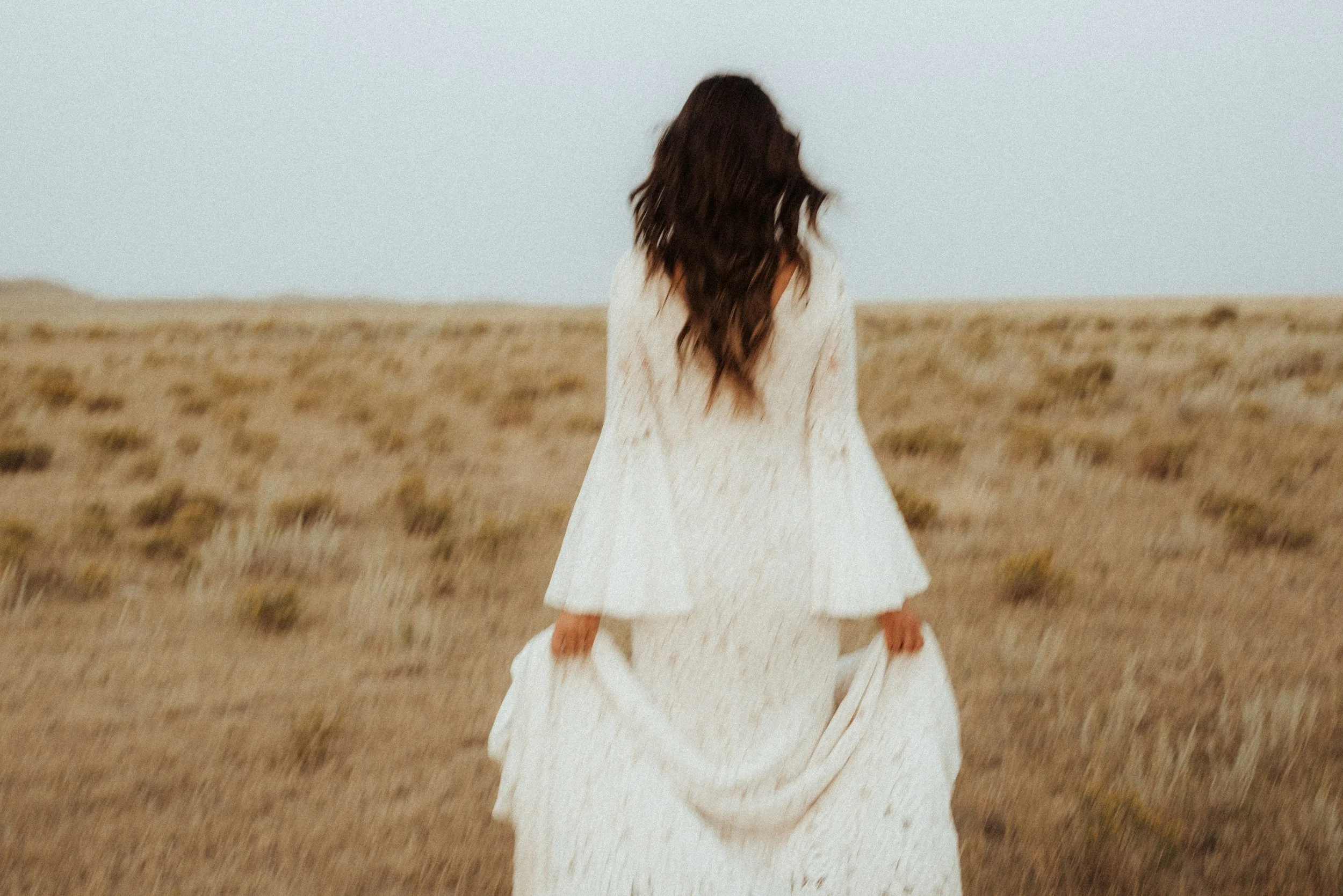 A woman with long, wavy dark hair walking away in a wide open desert landscape, wearing a flowing white dress.