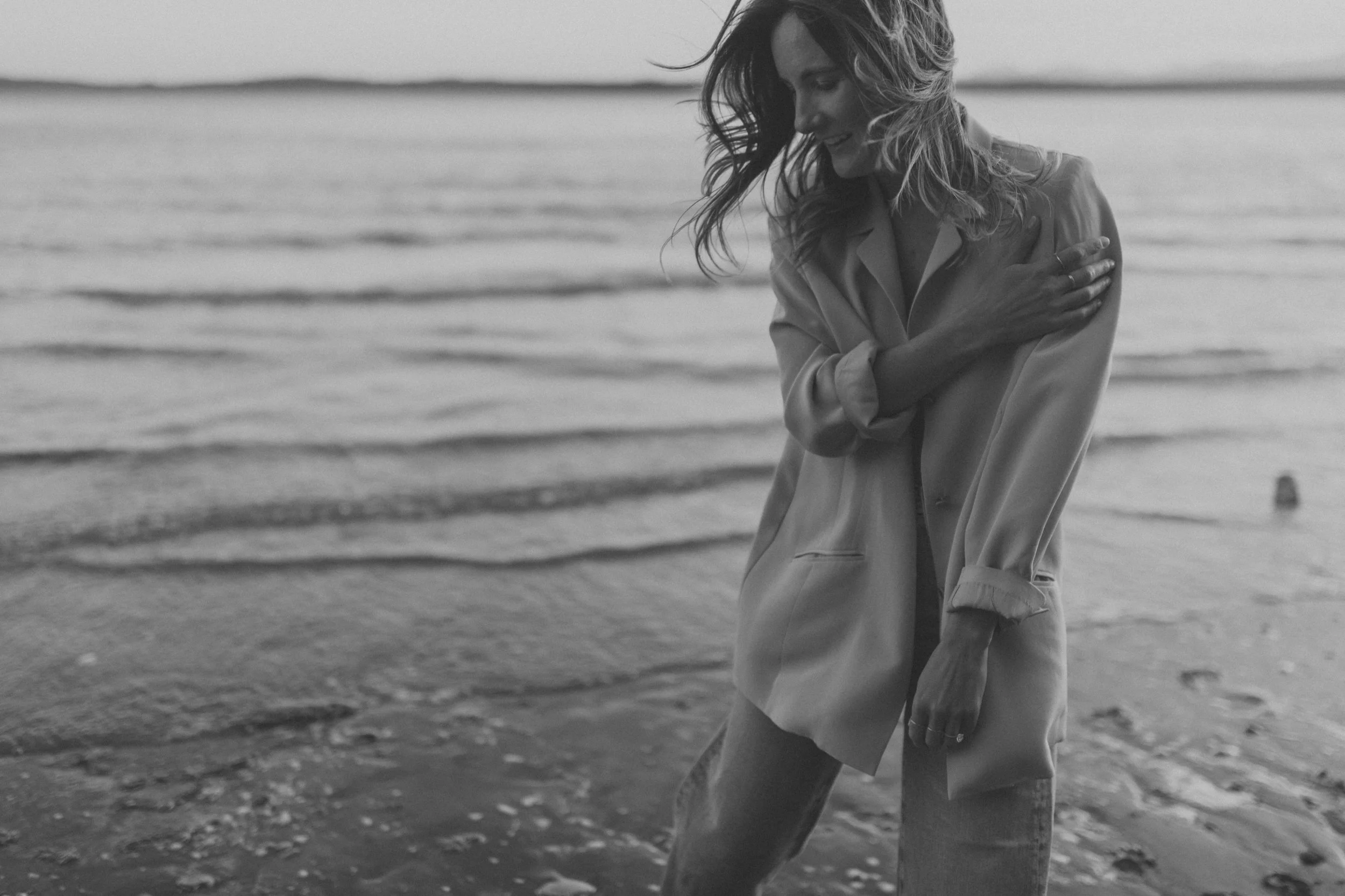 A woman standing on the beach with water and ripples in the background, smiling and hugging herself.