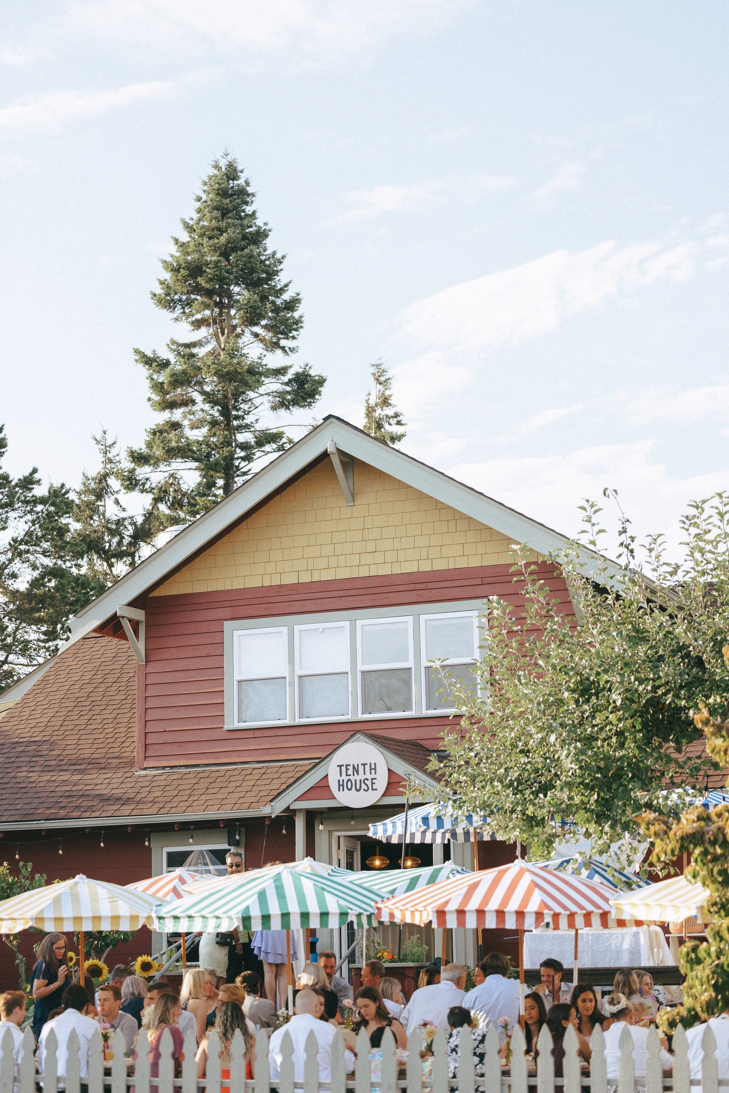 A gathering of people outside a restaurant named 'Tenth House' with colorful striped umbrellas, some people taking photos, and trees in the background.
