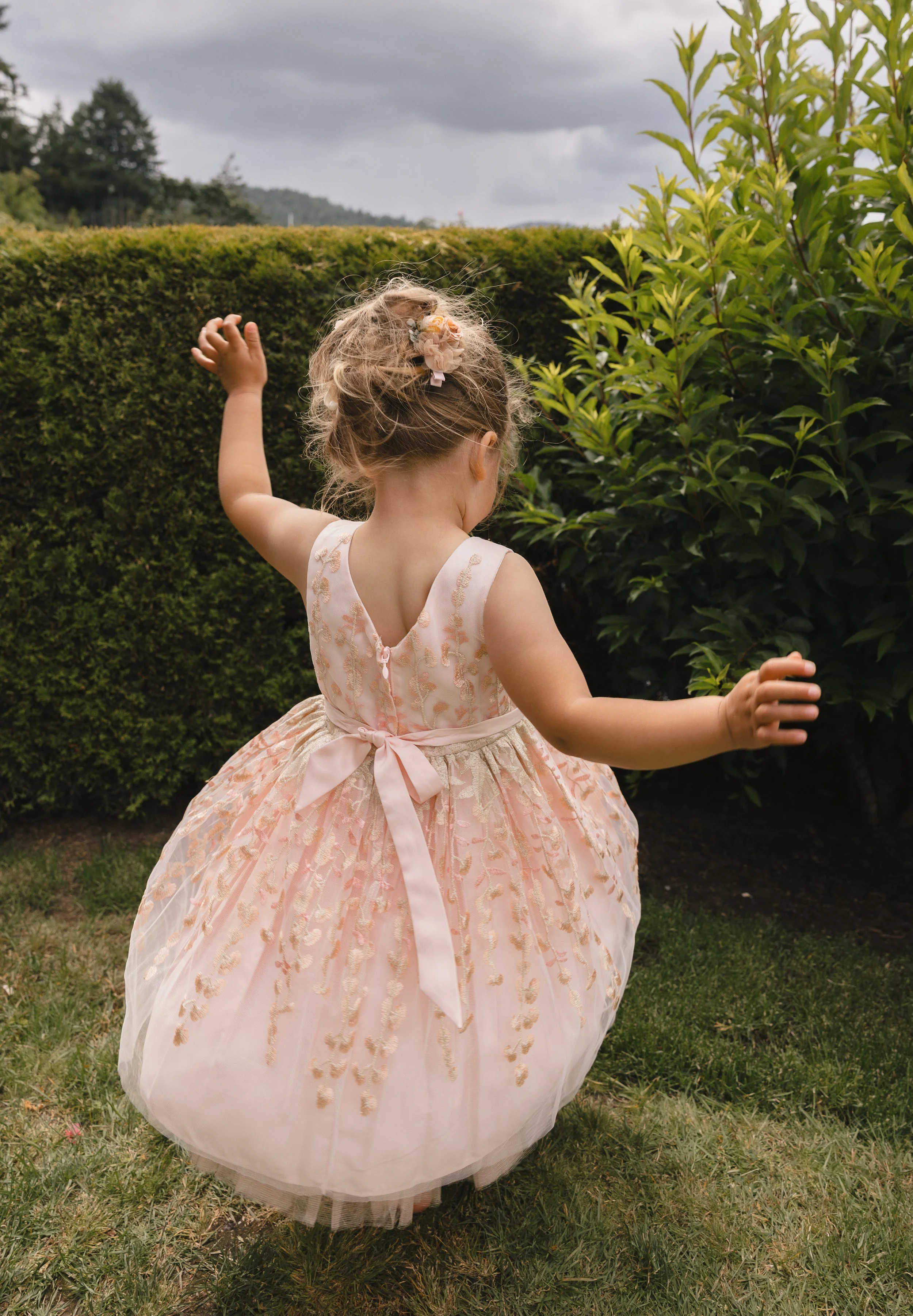 A young girl in a pink dress playing outdoors near bushes on a cloudy day.