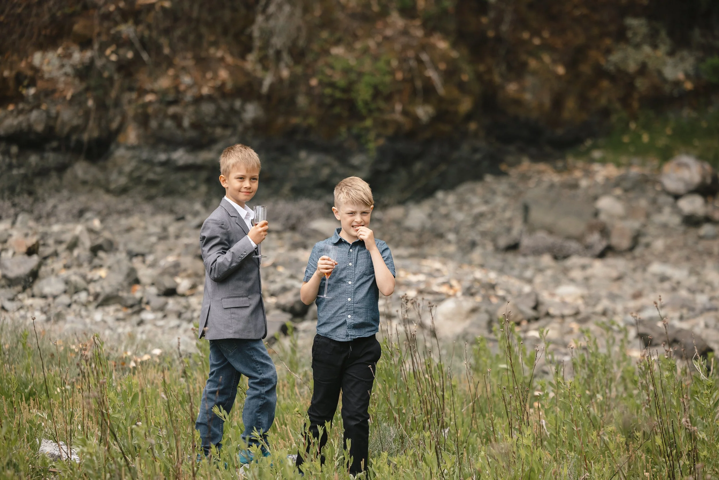 Two boys in formal attire holding drinks in a natural outdoor setting with rocky terrain and greenery.