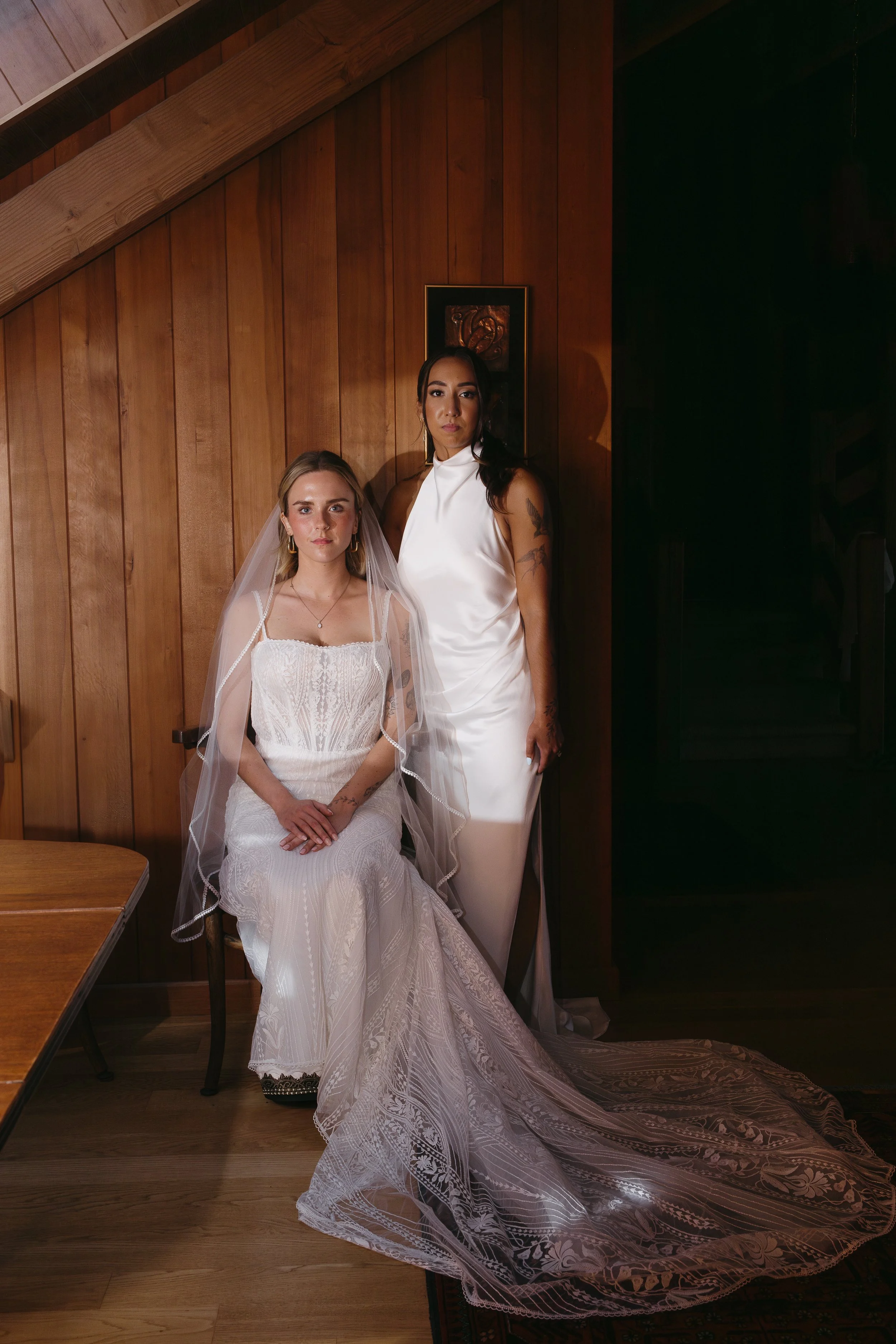 Two women in wedding attire inside a room with wooden walls. One is seated in a chair wearing a white wedding dress and veil, and the other stands beside her in a satin white gown.