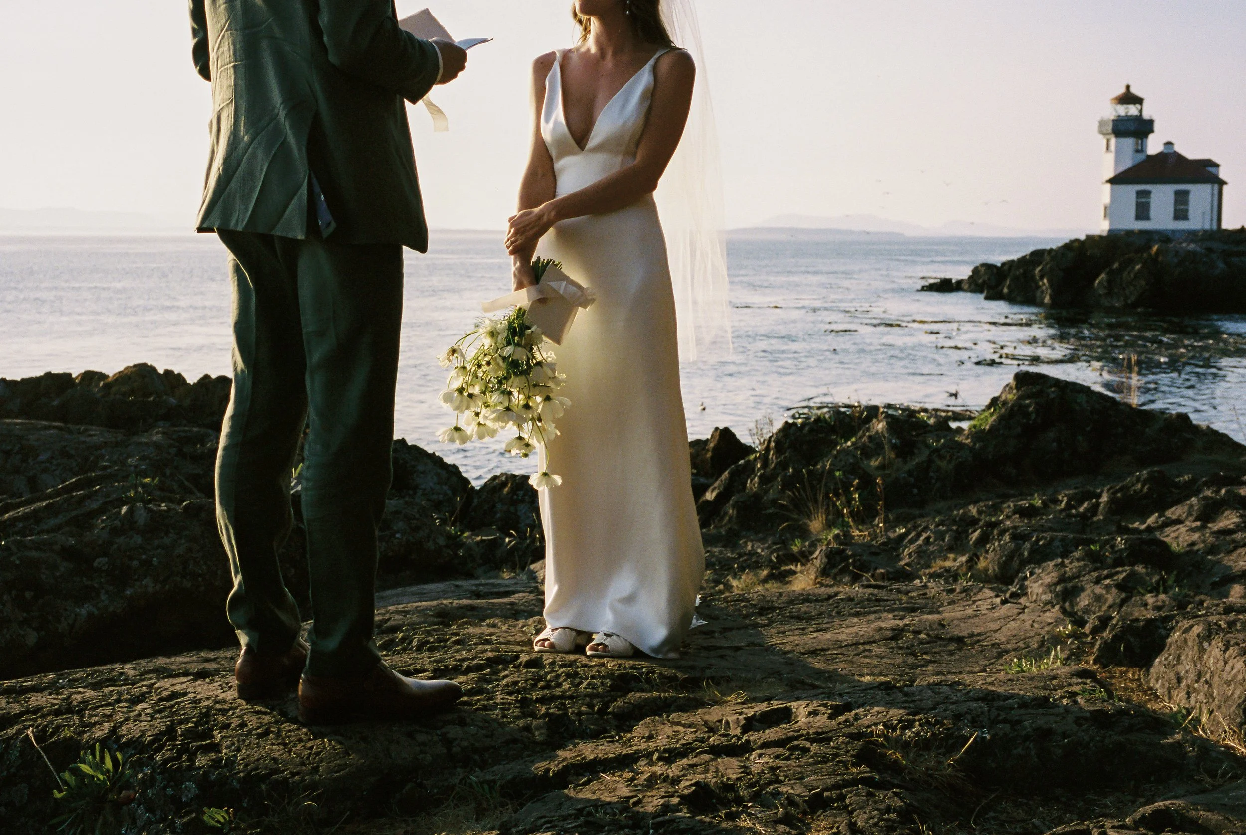 A couple gets married outdoors on a rocky shoreline at sunset, with a lighthouse in the background.
