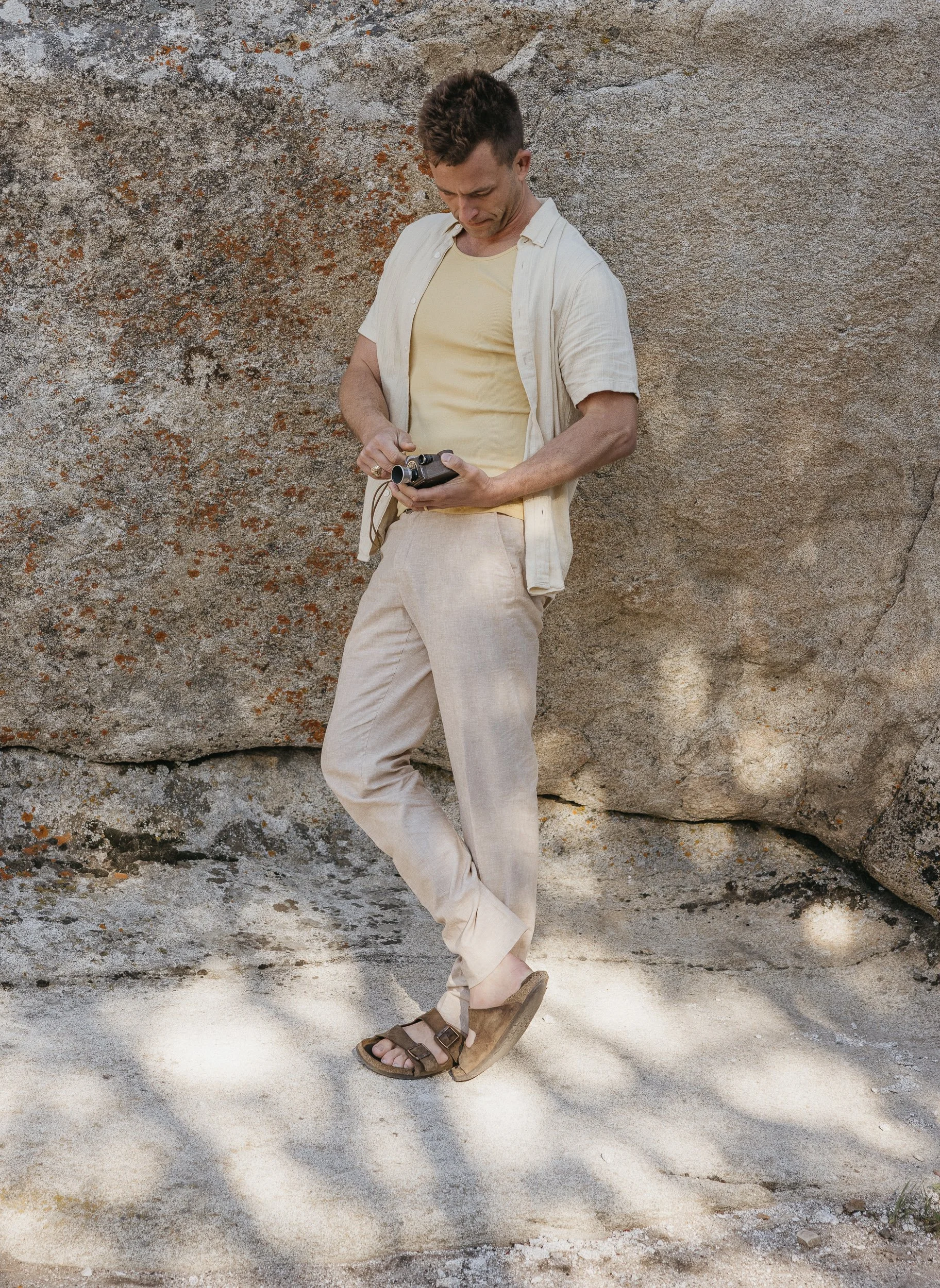 A man in beige pants, a yellow t-shirt, and a light-colored shirt stands against a large rock wall, looking at a remote control in his hands.