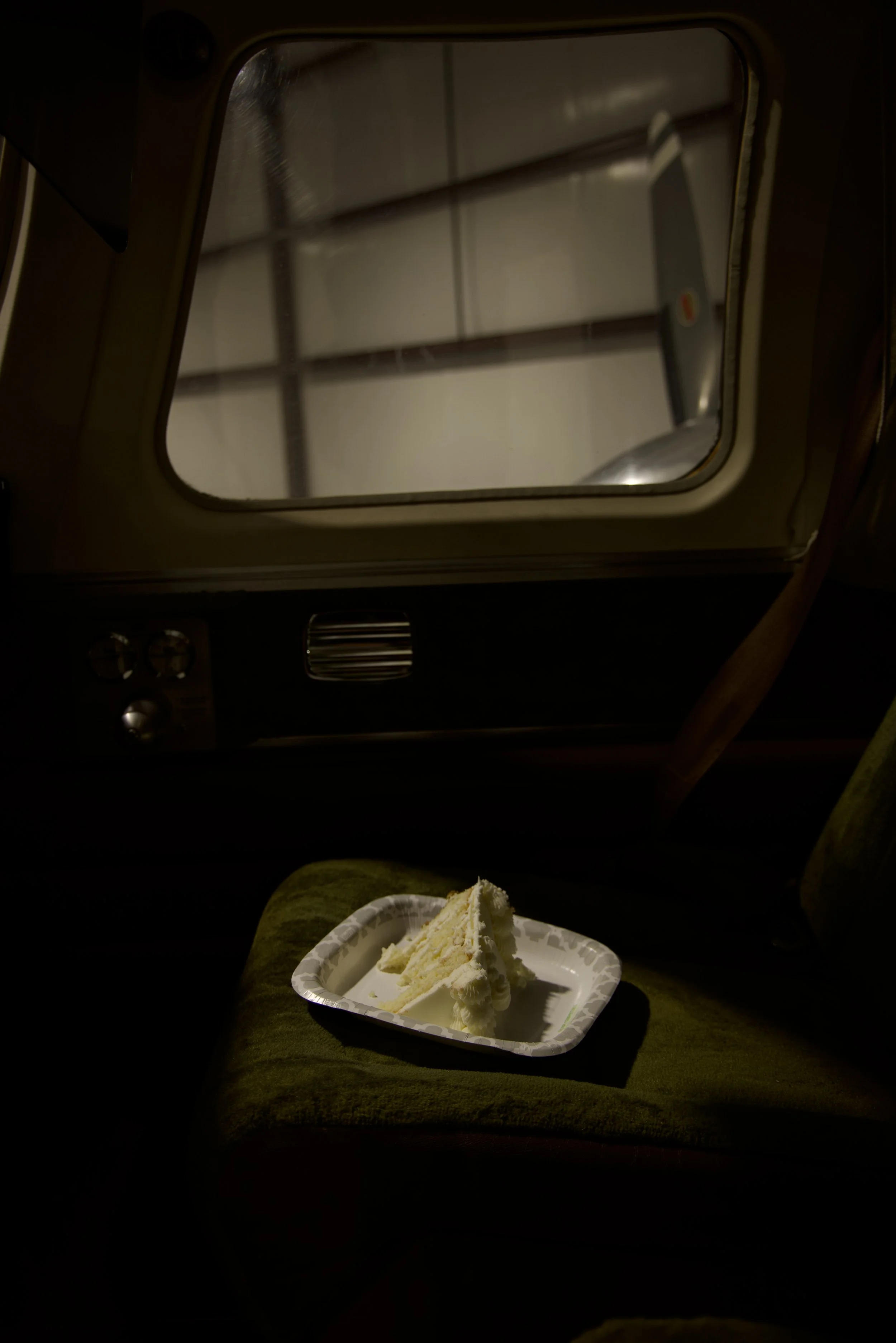 A piece of vanilla cake on a white disposable plate placed on a green seat, with a view of an airplane window showing the overcast sky outside.