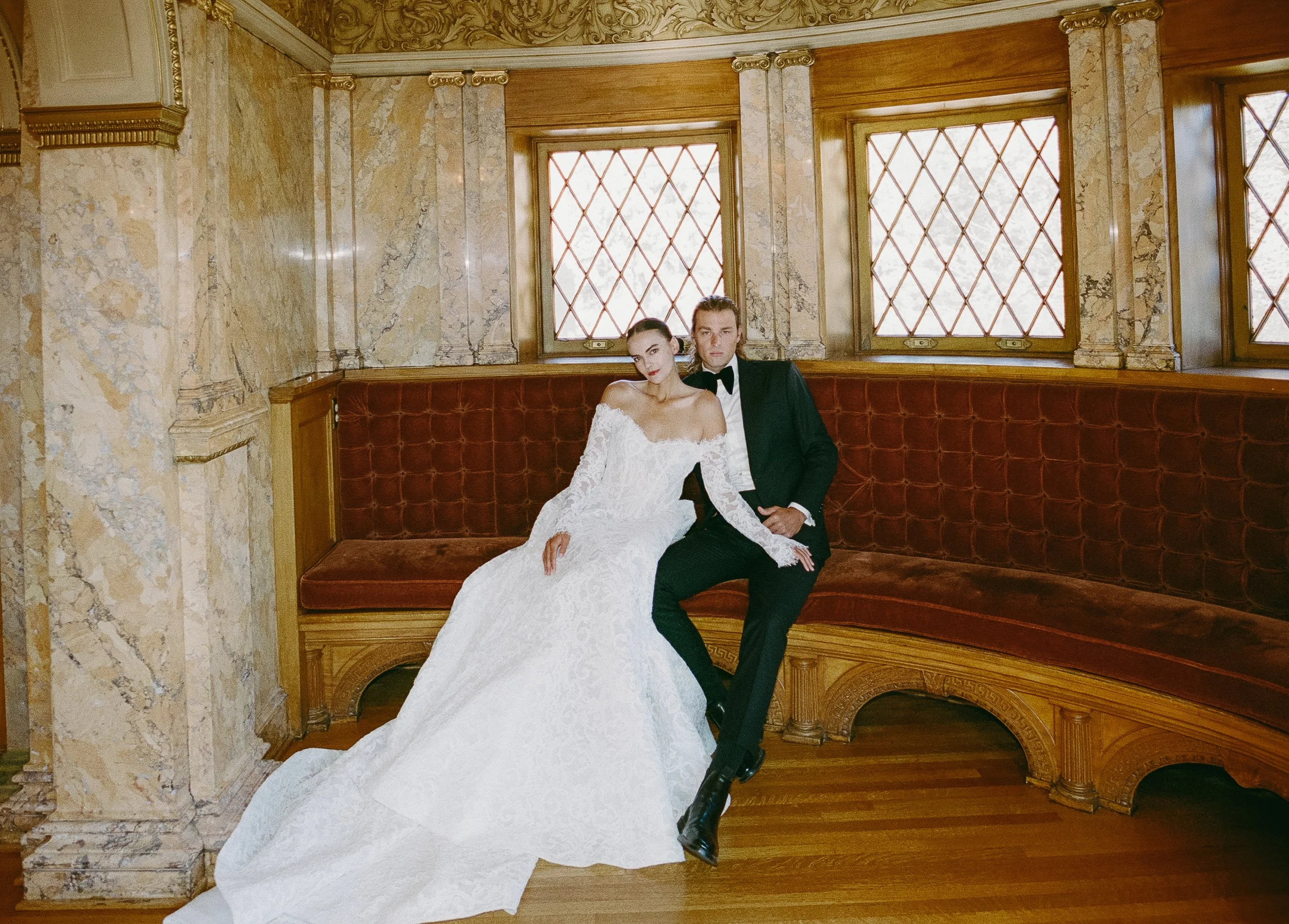 A bride and groom sitting on a curved red velvet bench inside a luxurious room with marble walls and wood accents. The bride is wearing a white off-shoulder wedding gown, and the groom is dressed in a black tuxedo with a bowtie.