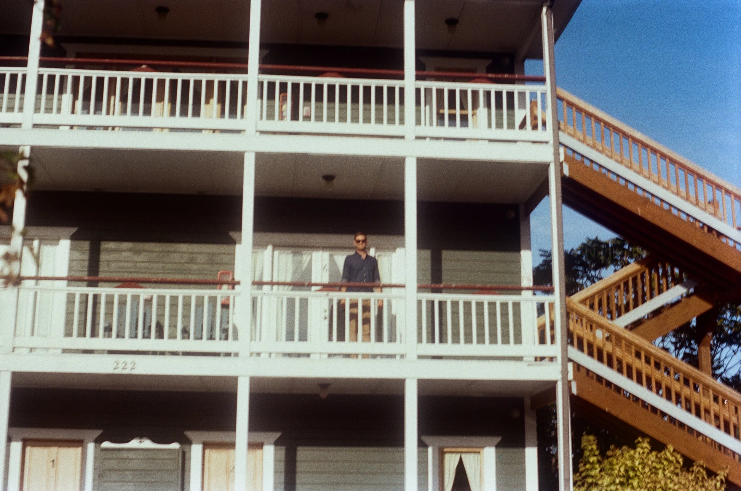 A man standing on an outdoor apartment balcony with white railings, with a wooden staircase to the right, under a clear blue sky.