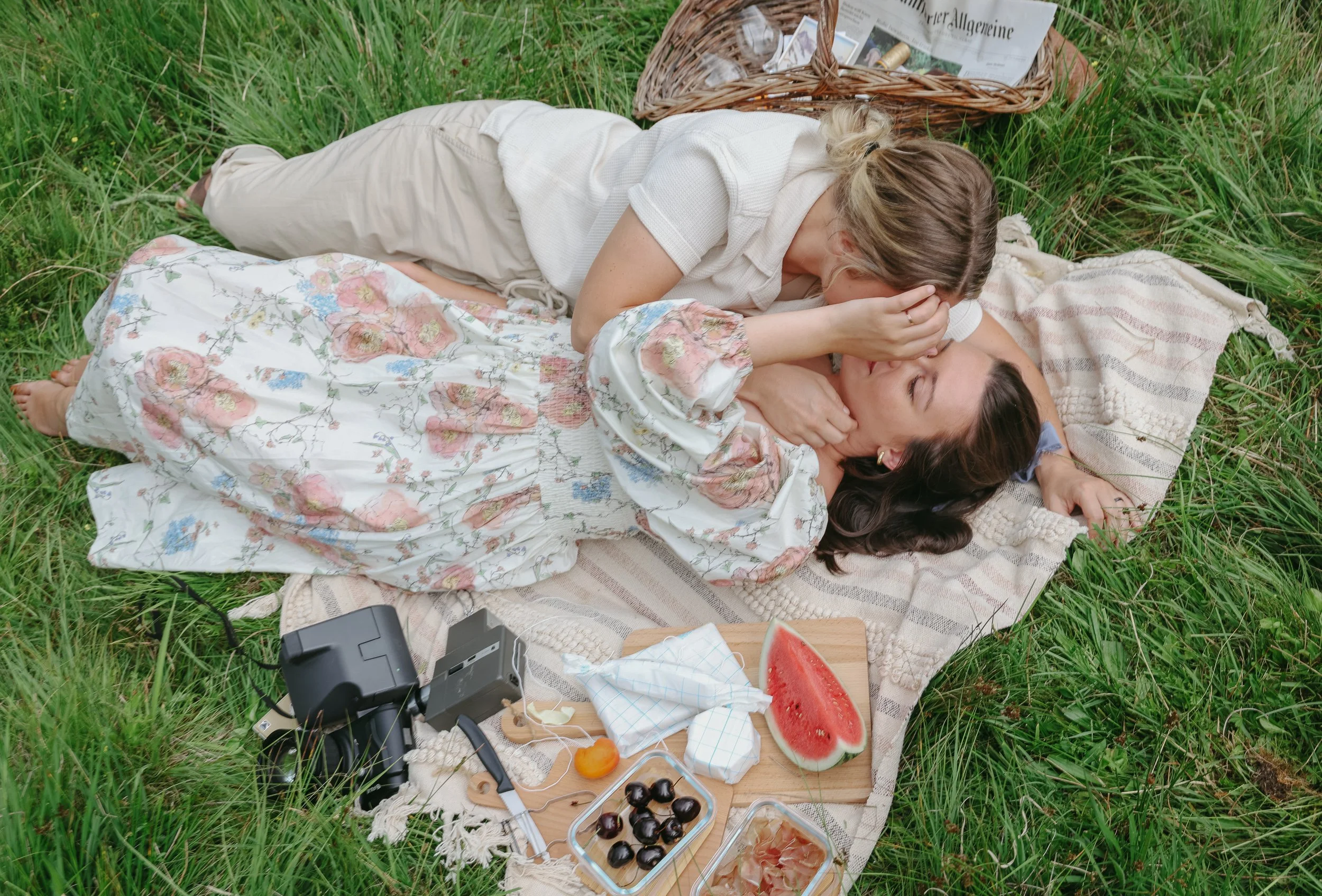 Two women on a picnic blanket in a grassy area, one woman lying and the other leaning over, as if kissing or whispering. There are food items including watermelon, cherries, and snacks, a camera, and picnic supplies surrounding them.