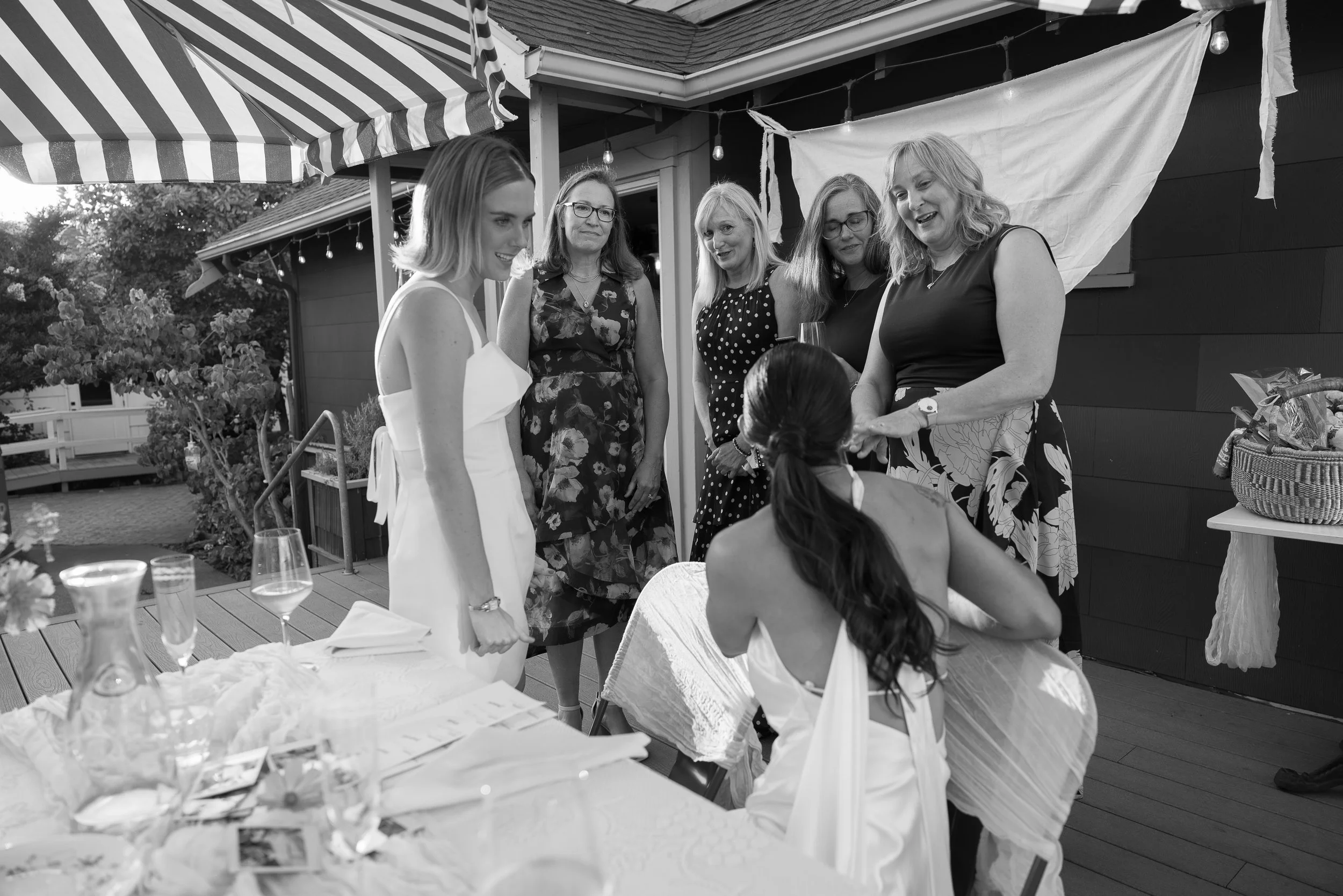 Group of women at a celebration, with a young woman in a white dress talking to a seated woman in a sleeveless dress, outdoors on a decorated patio.