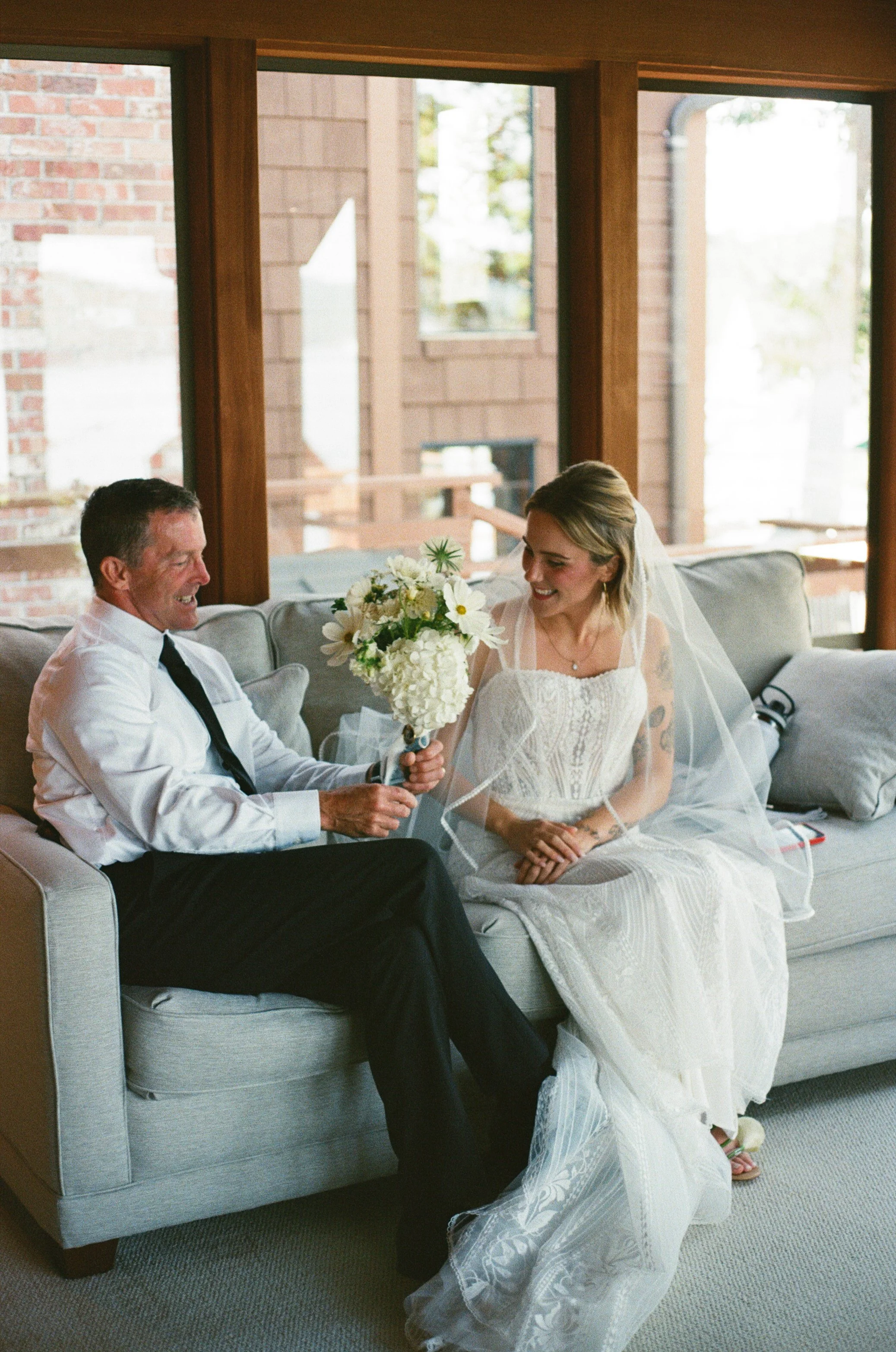 A bride and an older man, possibly her father, sitting on a light-colored couch inside a room with large windows. The man is giving the bride a bouquet of white and yellow flowers. The bride is wearing a white wedding dress with a lace design and a veil. Both are smiling, sharing a joyful moment.