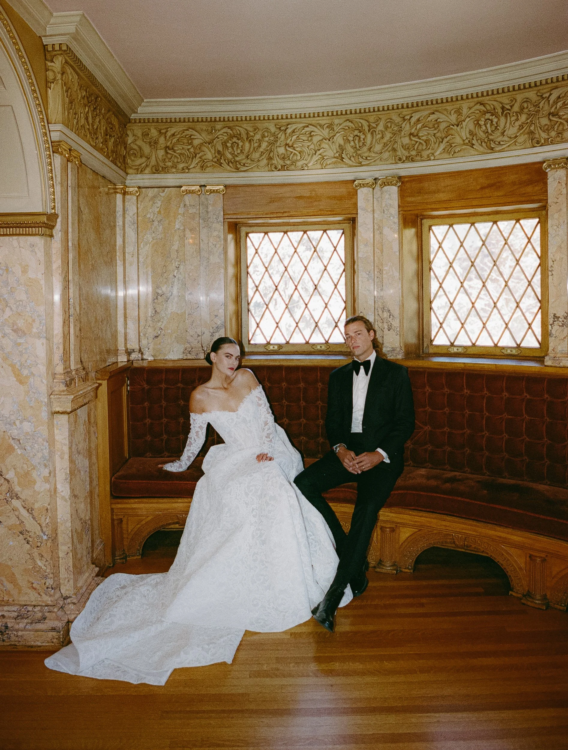Elegant couple in wedding attire sitting on a curved velvet bench in a historic, ornately decorated room with large patterned windows and marble walls.