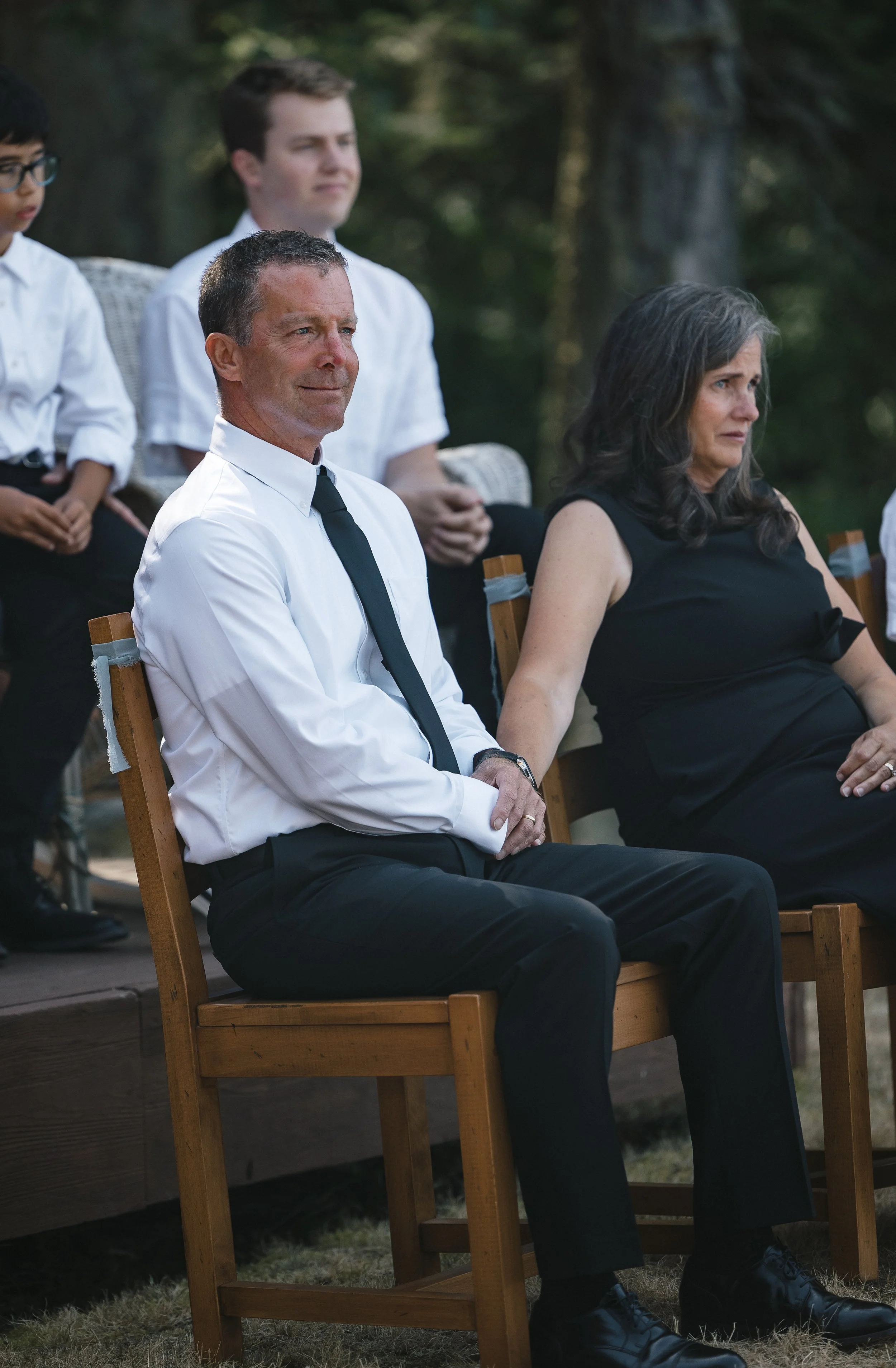 A man and a woman sitting on chairs outdoors at a formal event, with other people standing behind them.