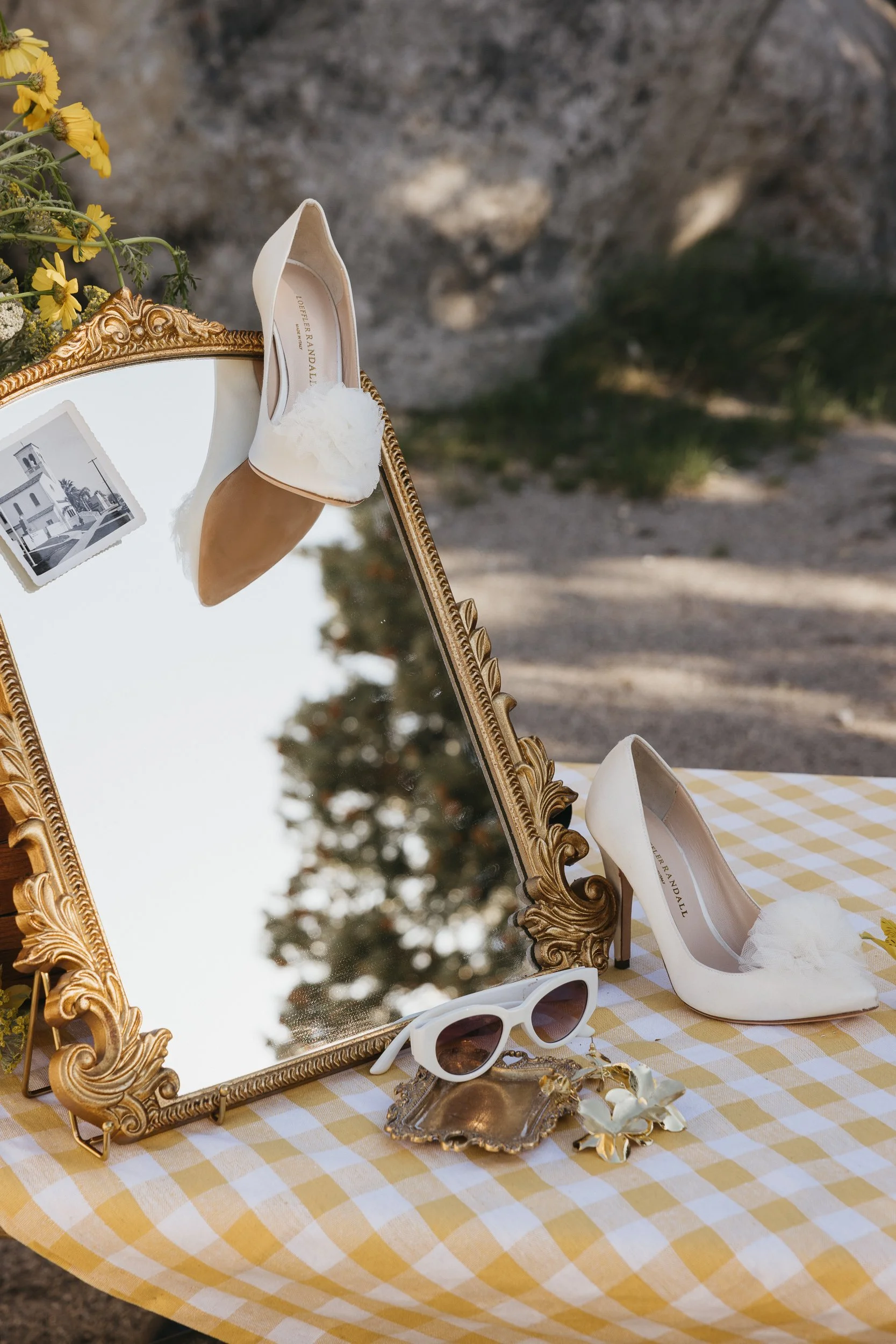White high-heeled shoes with tulle embellishment, white sunglasses, and small decorative jewelry on a yellow and white checkered tablecloth, with a gold ornate mirror reflecting the shoes, outdoors with a gravel ground and some greenery in the background.