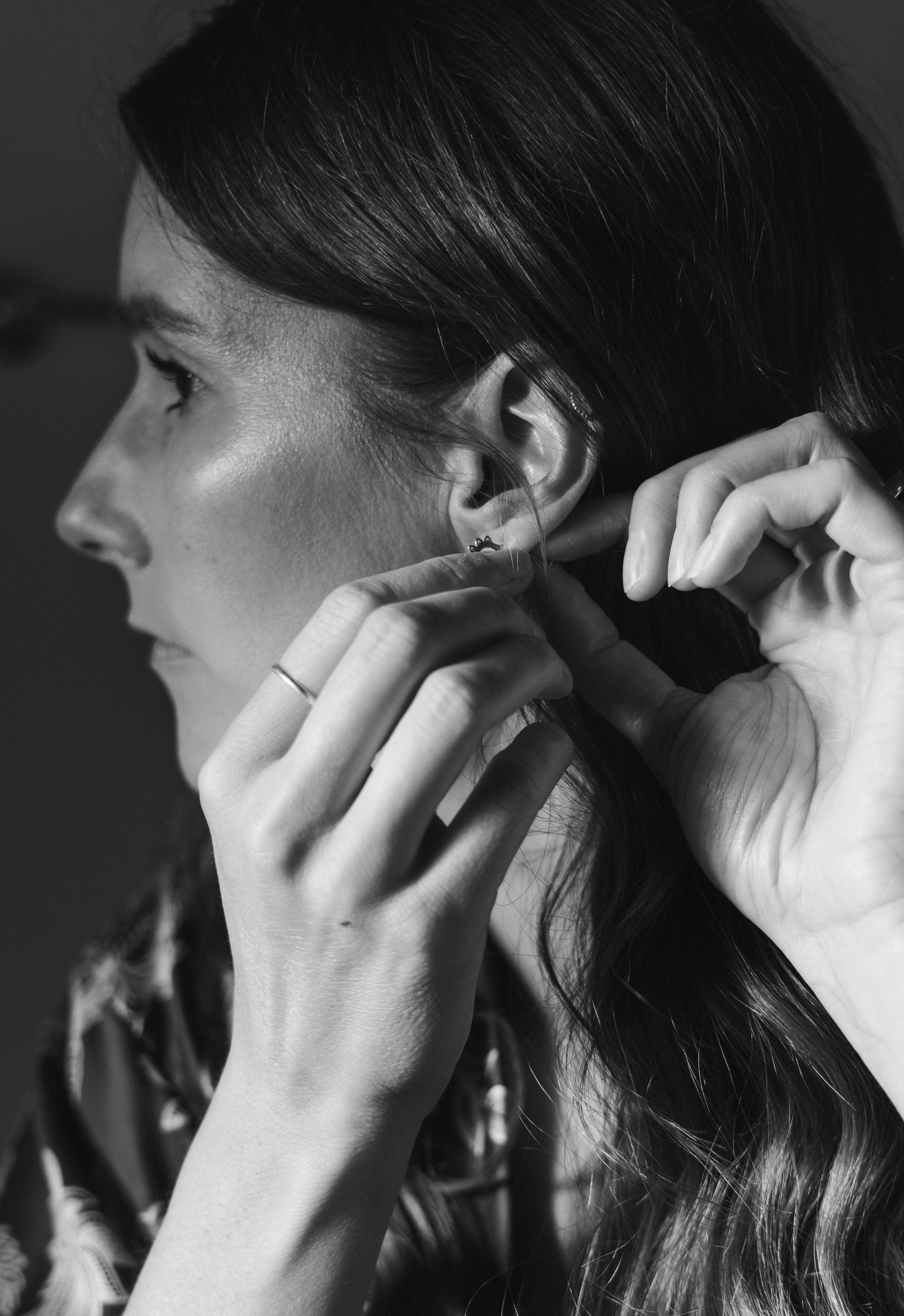 A woman with dark hair is putting on an earring in her ear, with her hands near her face, in a black-and-white photograph.