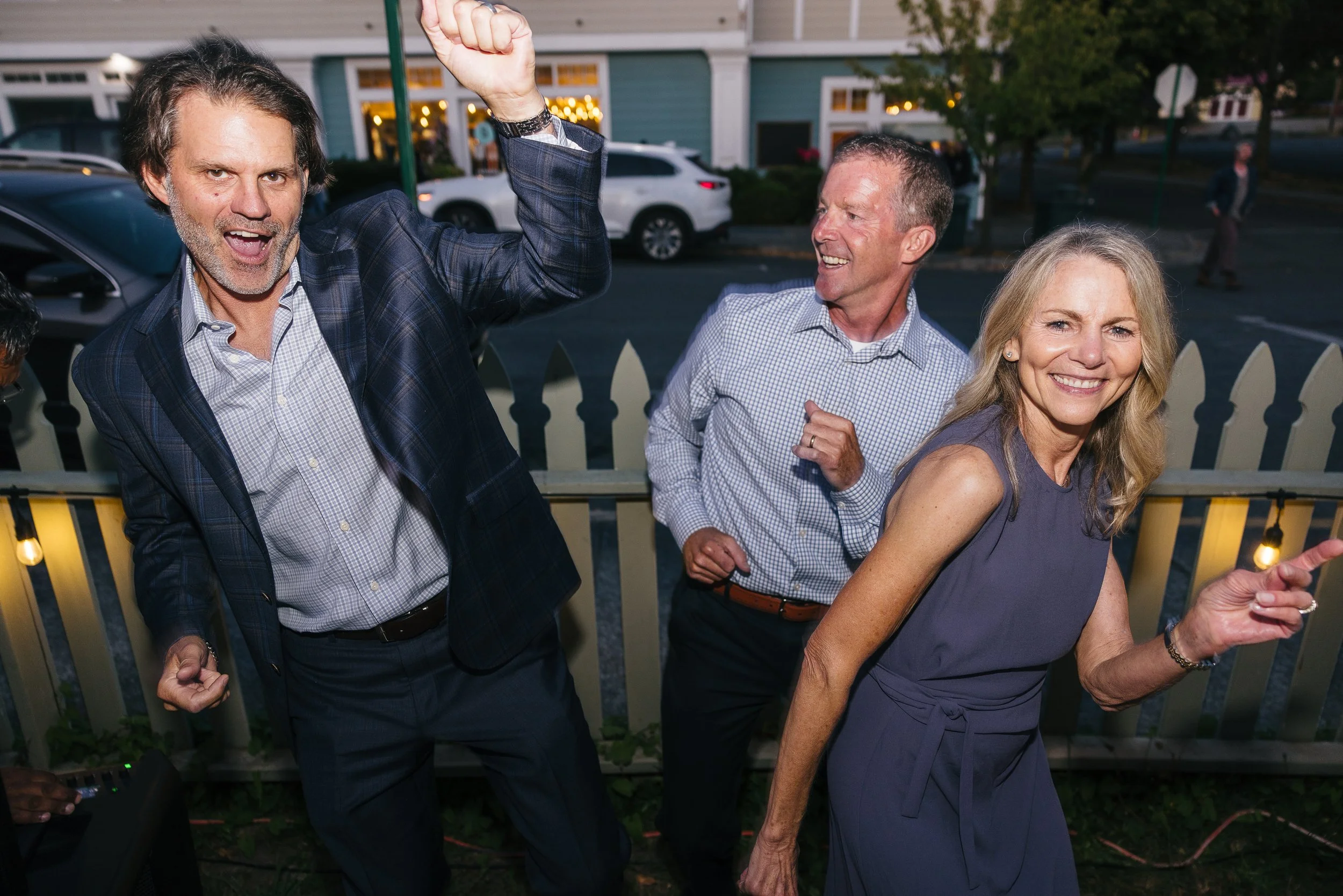 Four people at an outdoor gathering dancing and smiling, with a fence and houses in the background during evening.