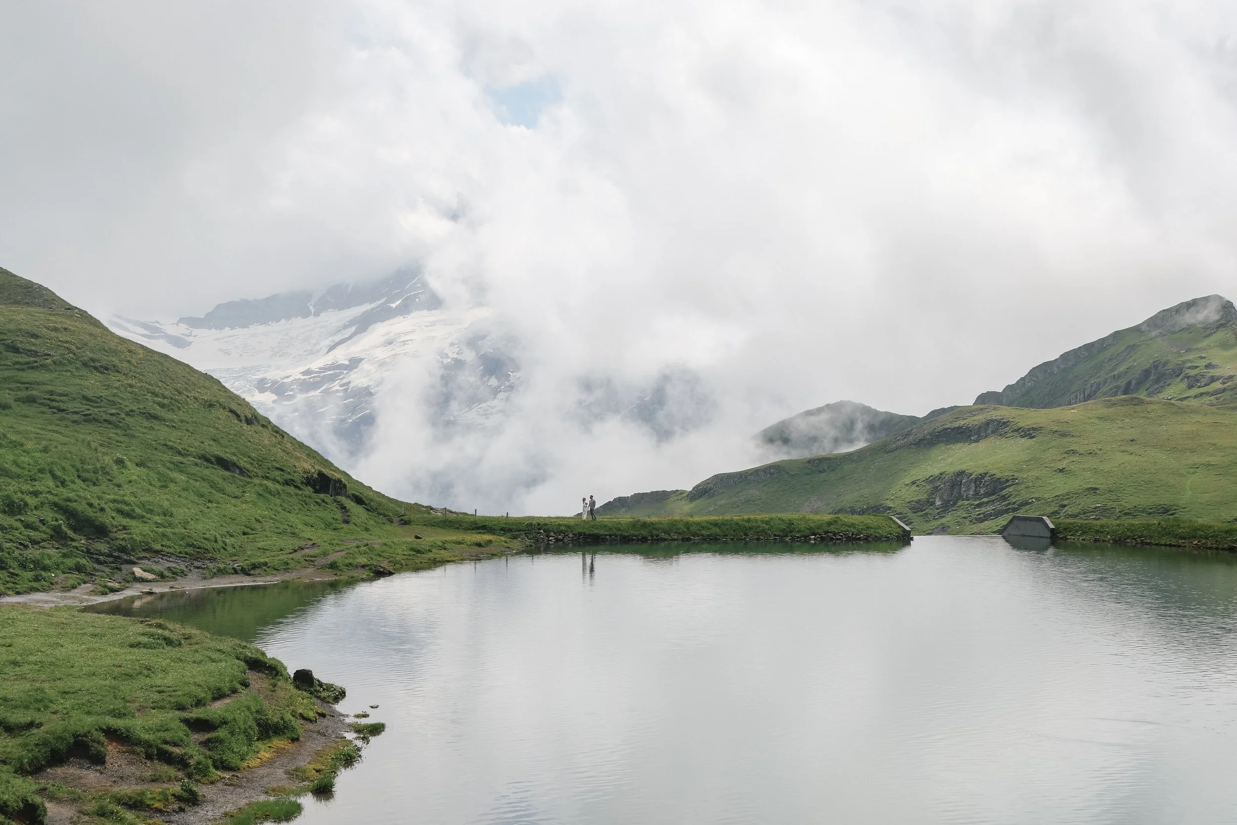 Mountain landscape with green hills, a small lake, and a cloudy sky.