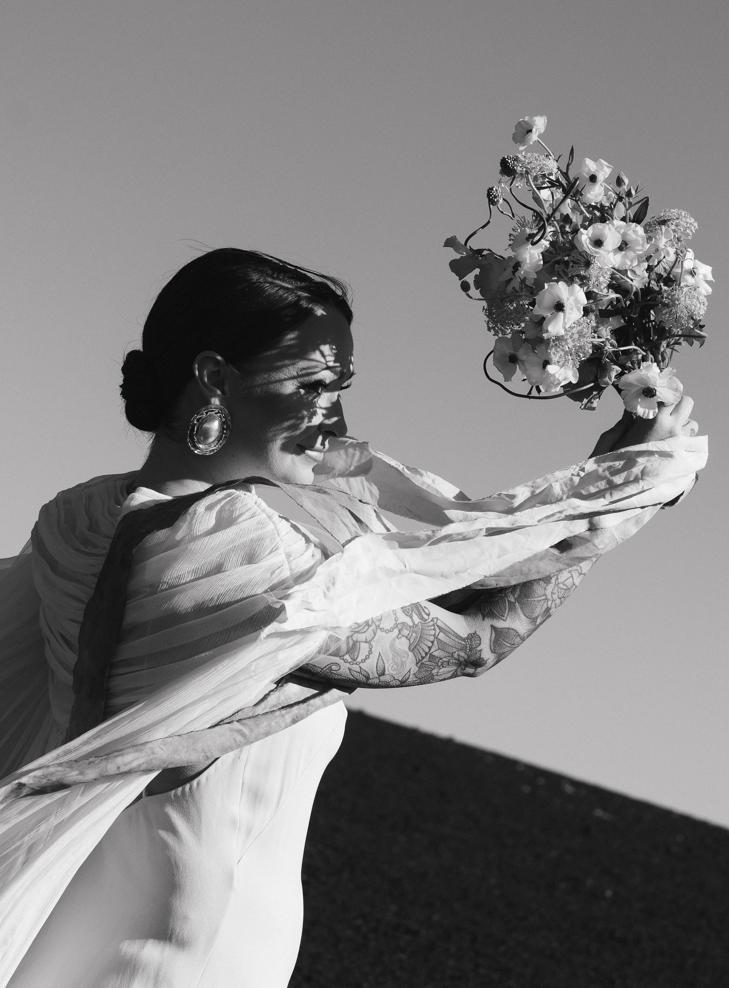 A woman dressed in a flowing outfit holding a bouquet of flowers, with shadows cast on her face from the branches, against a clear sky.