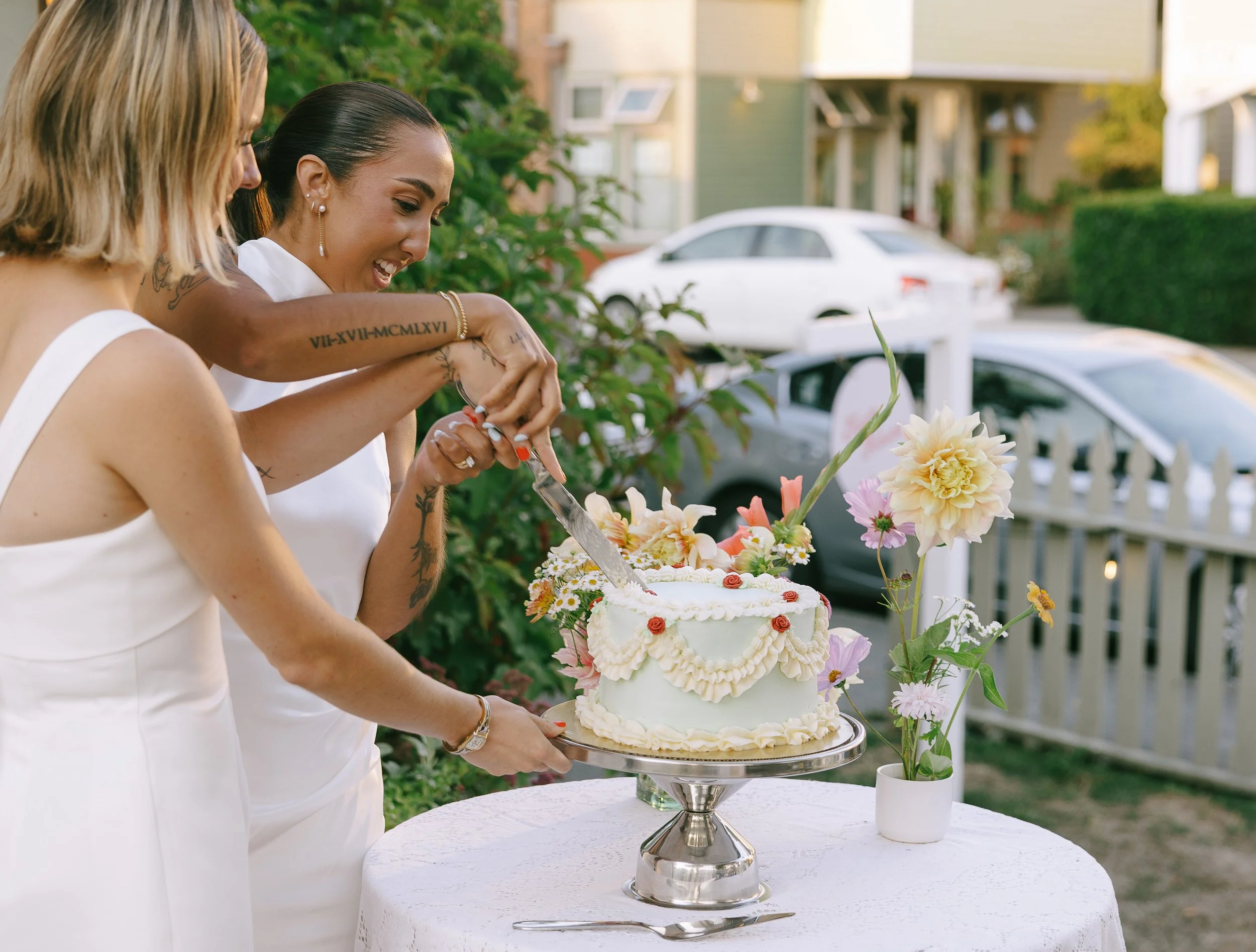 Two women in white dresses cut a wedding cake outdoors, with flowers and cars in the background.