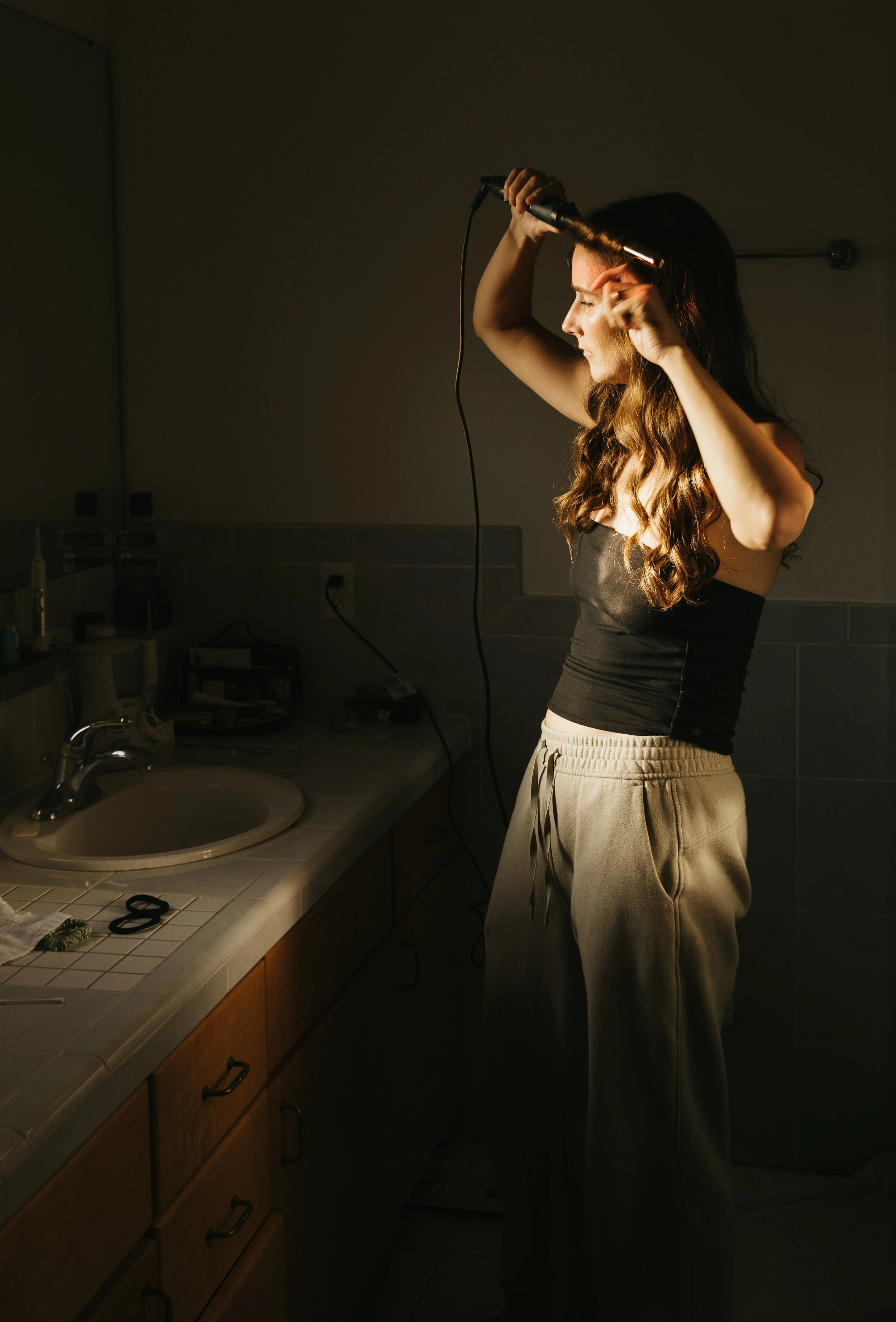 A woman with long, wavy brown hair straightening her hair with a curling iron in a bathroom, which has a sink, countertop, and various toiletries.