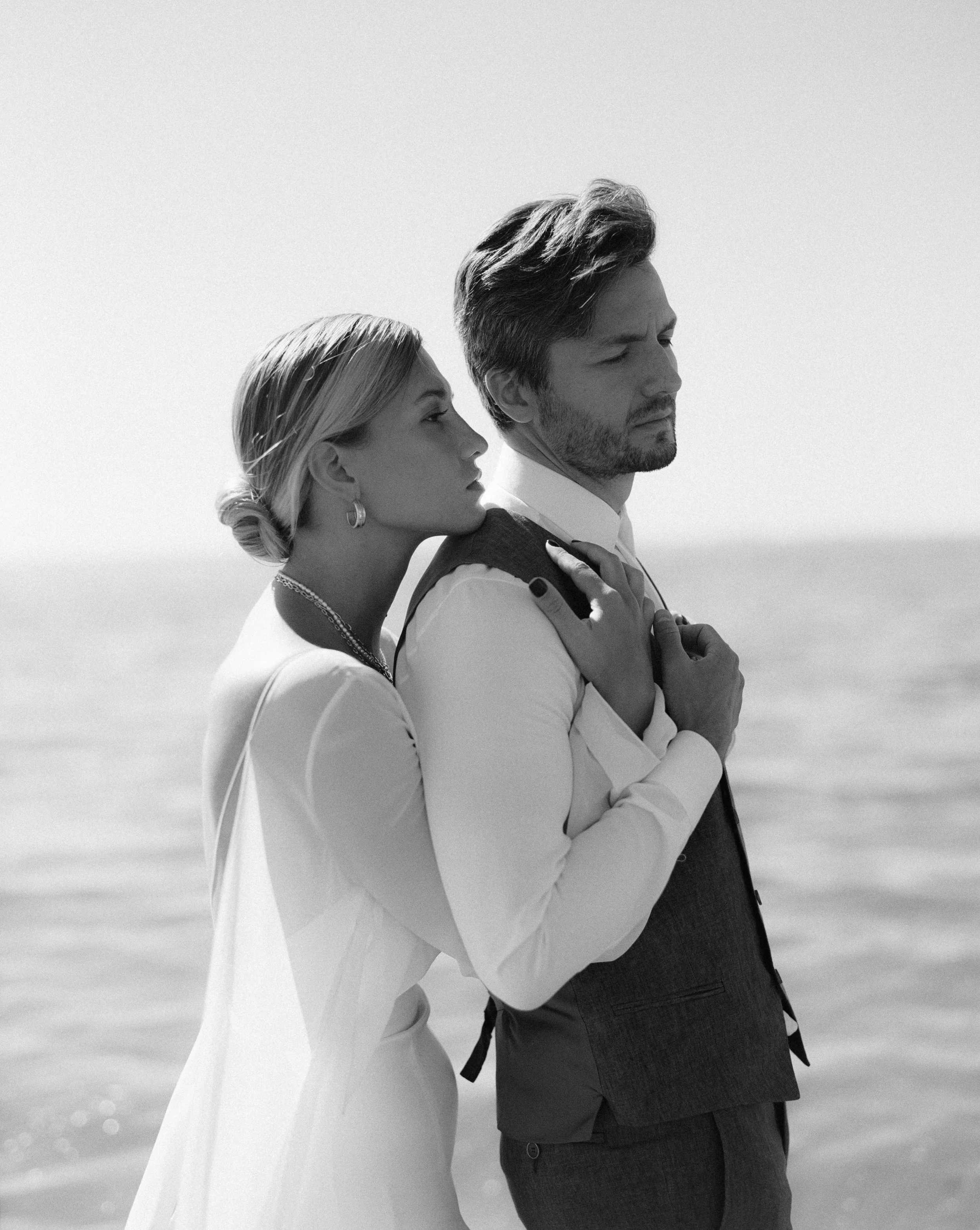 Black and white photo of a couple standing close together near water, with the woman leaning on the man's shoulder. The woman has styled hair, earrings, and a necklace, and is wearing a white dress. The man has styled hair, a beard, and is dressed in a suit with a vest and a tie.